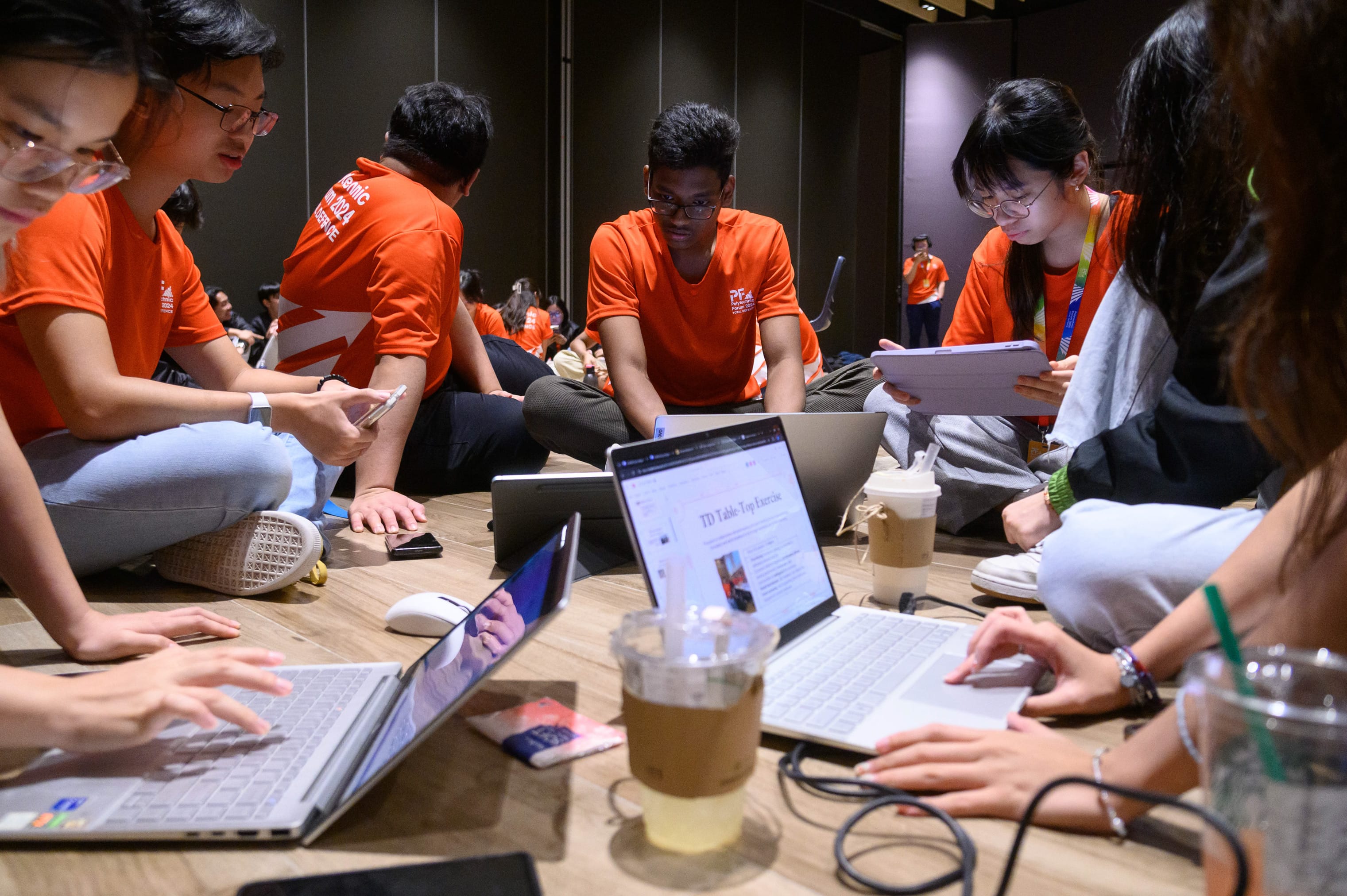 Several young people in orange shirts sit on the floor around laptops and drinks, working together on a group exercise.