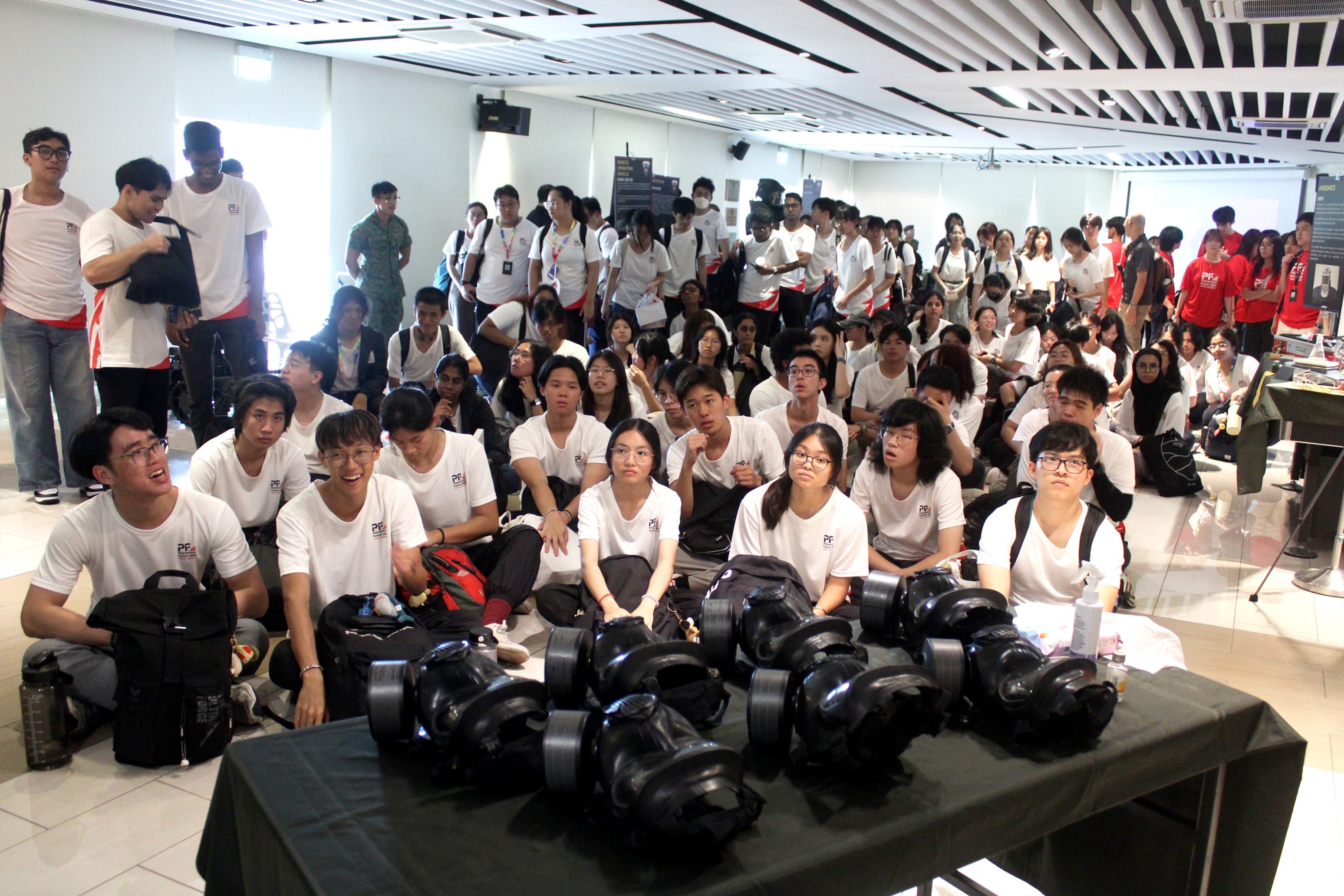Large group of young people in matching white t-shirts sitting and standing in a bright indoor hall.