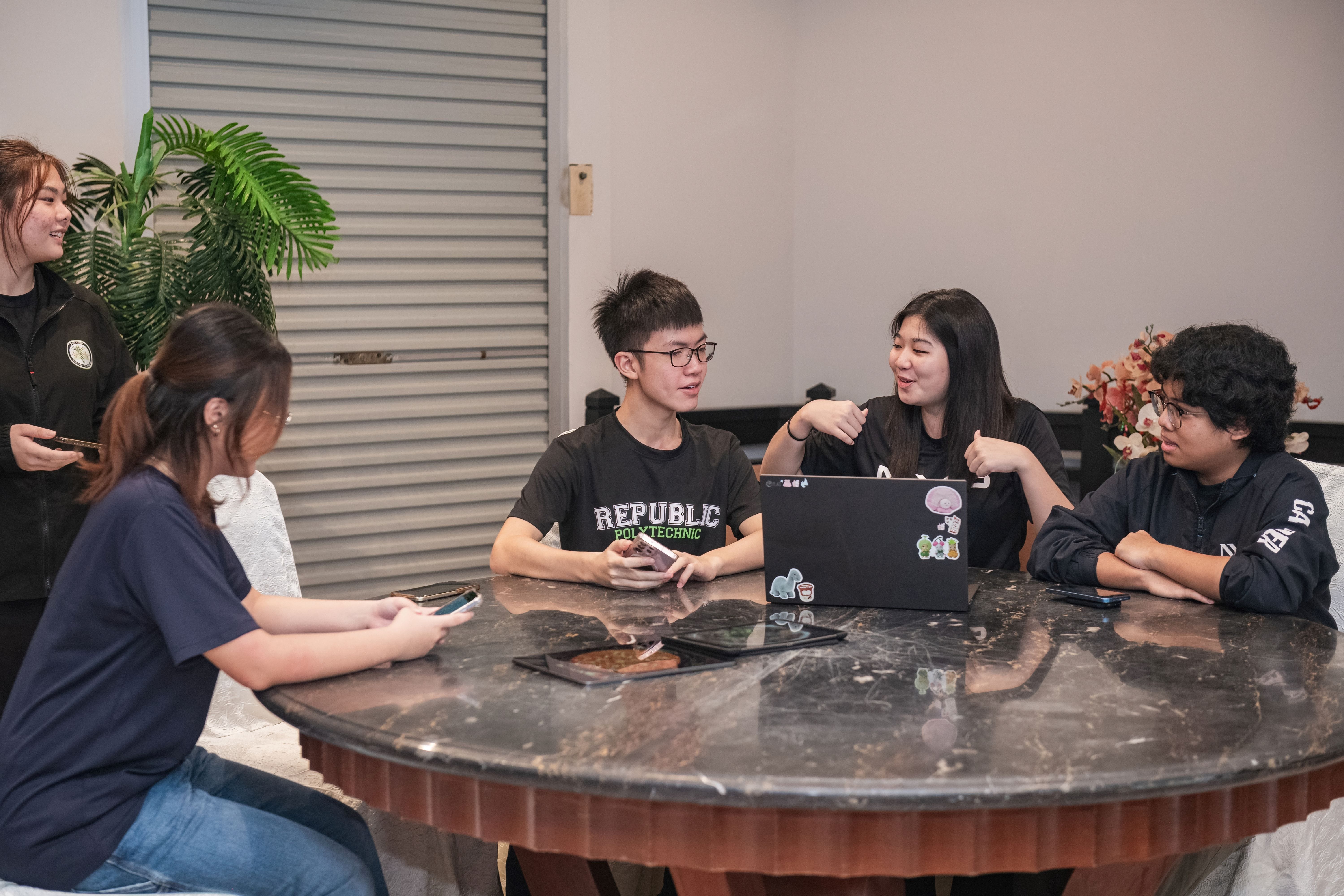 Five young people sit and stand around a round marble table with a sticker-covered laptop, talking and holding phones.