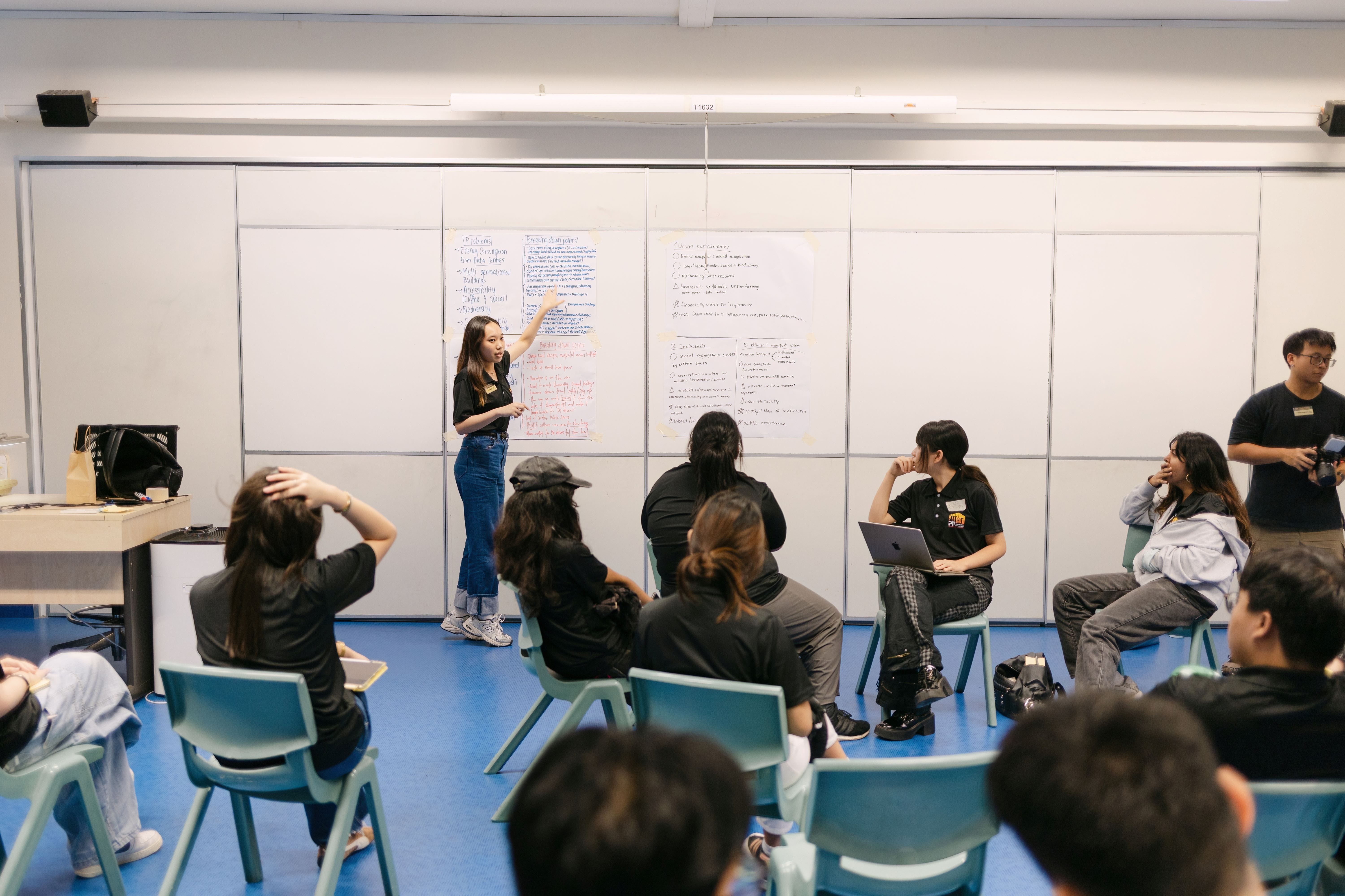 A young woman presents notes on large paper sheets mounted on a wall to a group of seated students in a classroom.