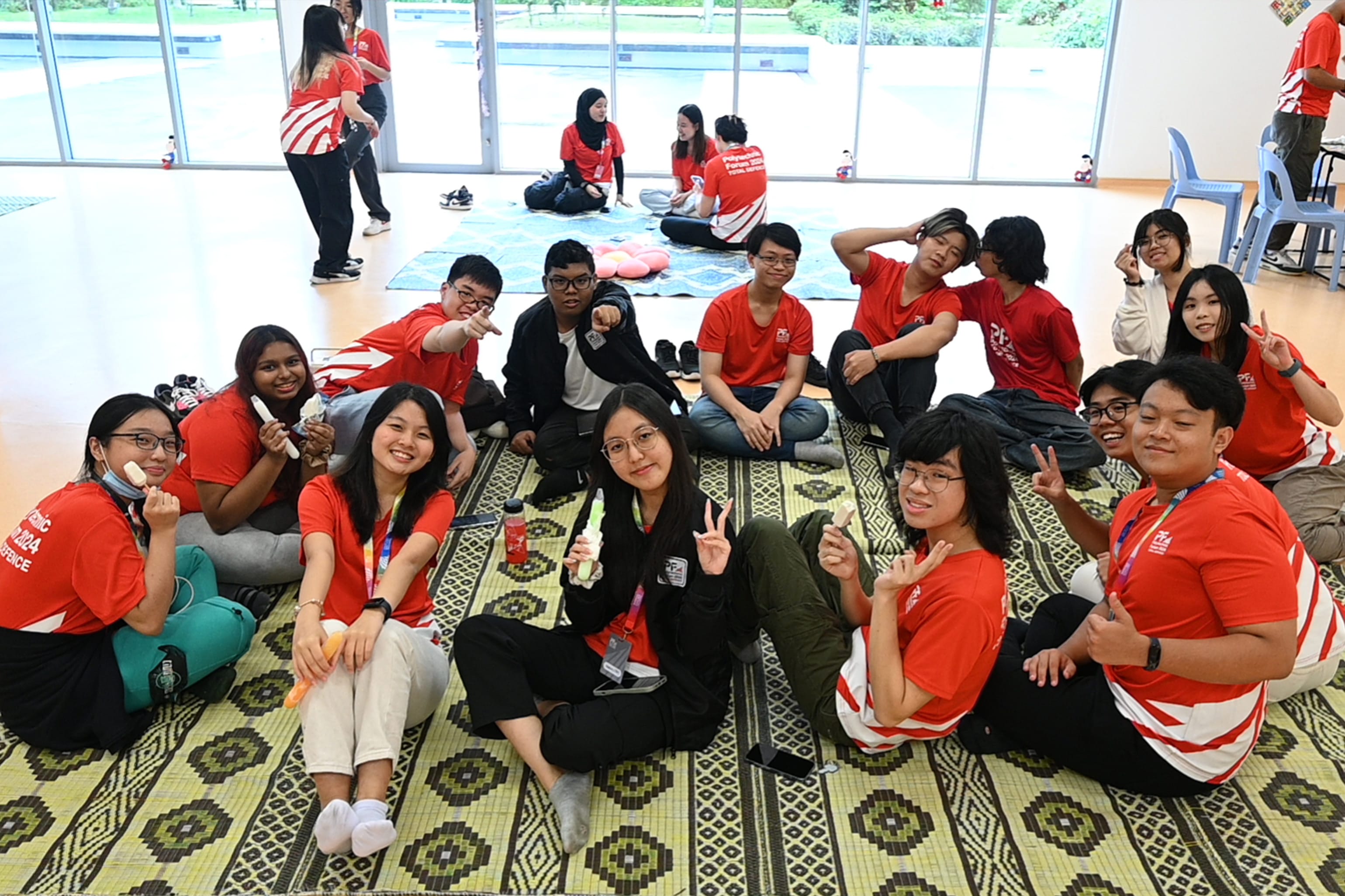 A large group of young people in red shirts sit on patterned mats indoors, smiling and posing for the camera.