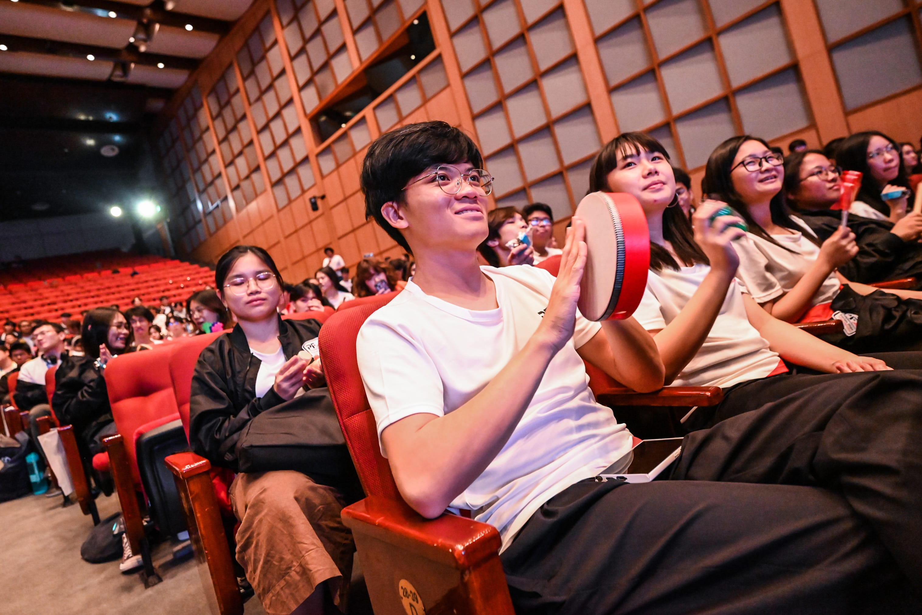 Young audience members sit in red theater seats, smiling and holding tambourines during a live event.
