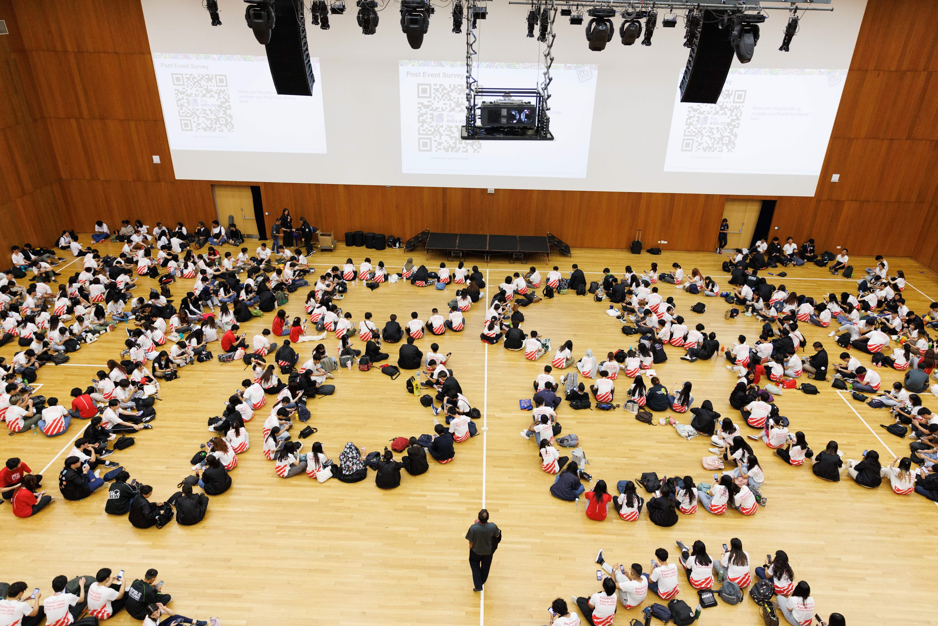 Large group of people in white shirts sitting on a gymnasium floor facing screens displaying QR codes.