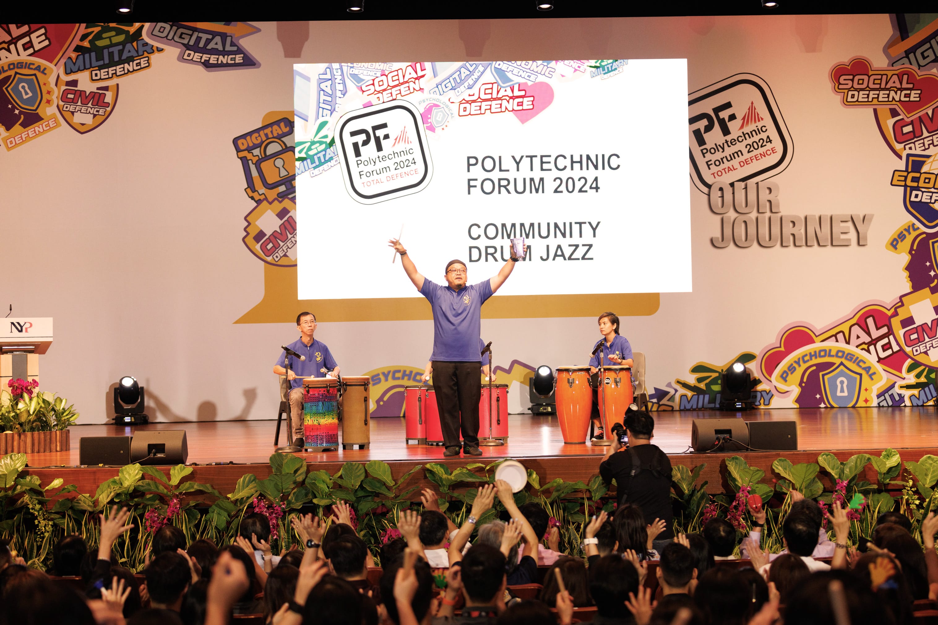 Three performers in blue shirts play drums on stage at Polytechnic Forum 2024 Community Drum Jazz event.