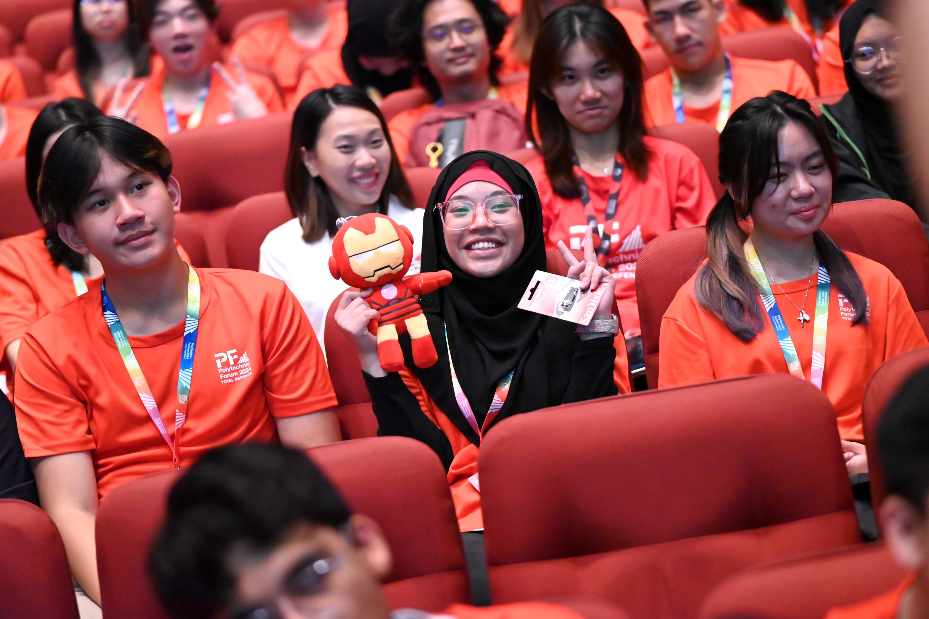A smiling woman in a black hijab holds an Iron Man plush toy while seated among students in red shirts.