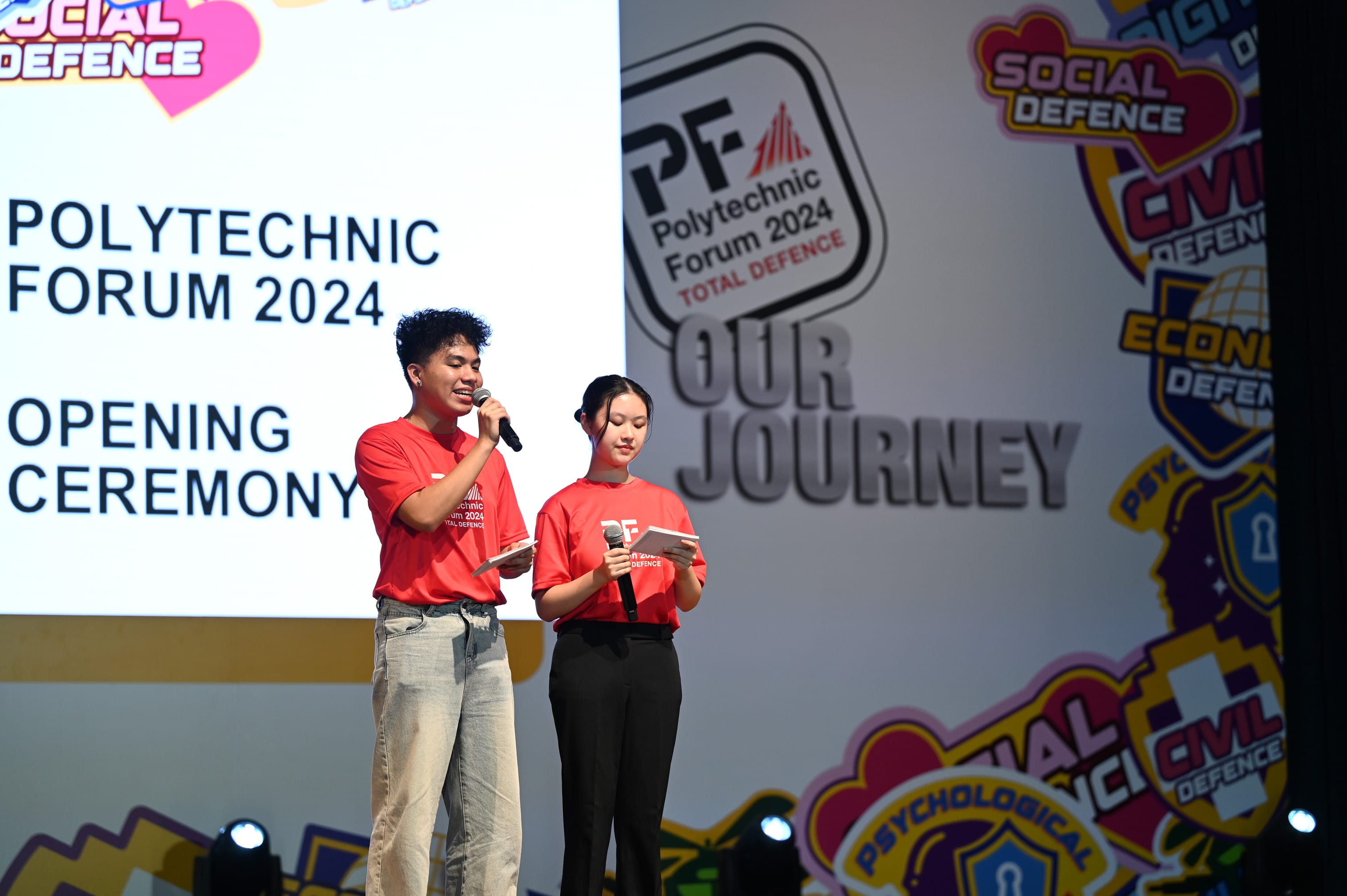 Two emcees in red shirts hold microphones on stage at the Polytechnic Forum 2024 Opening Ceremony.