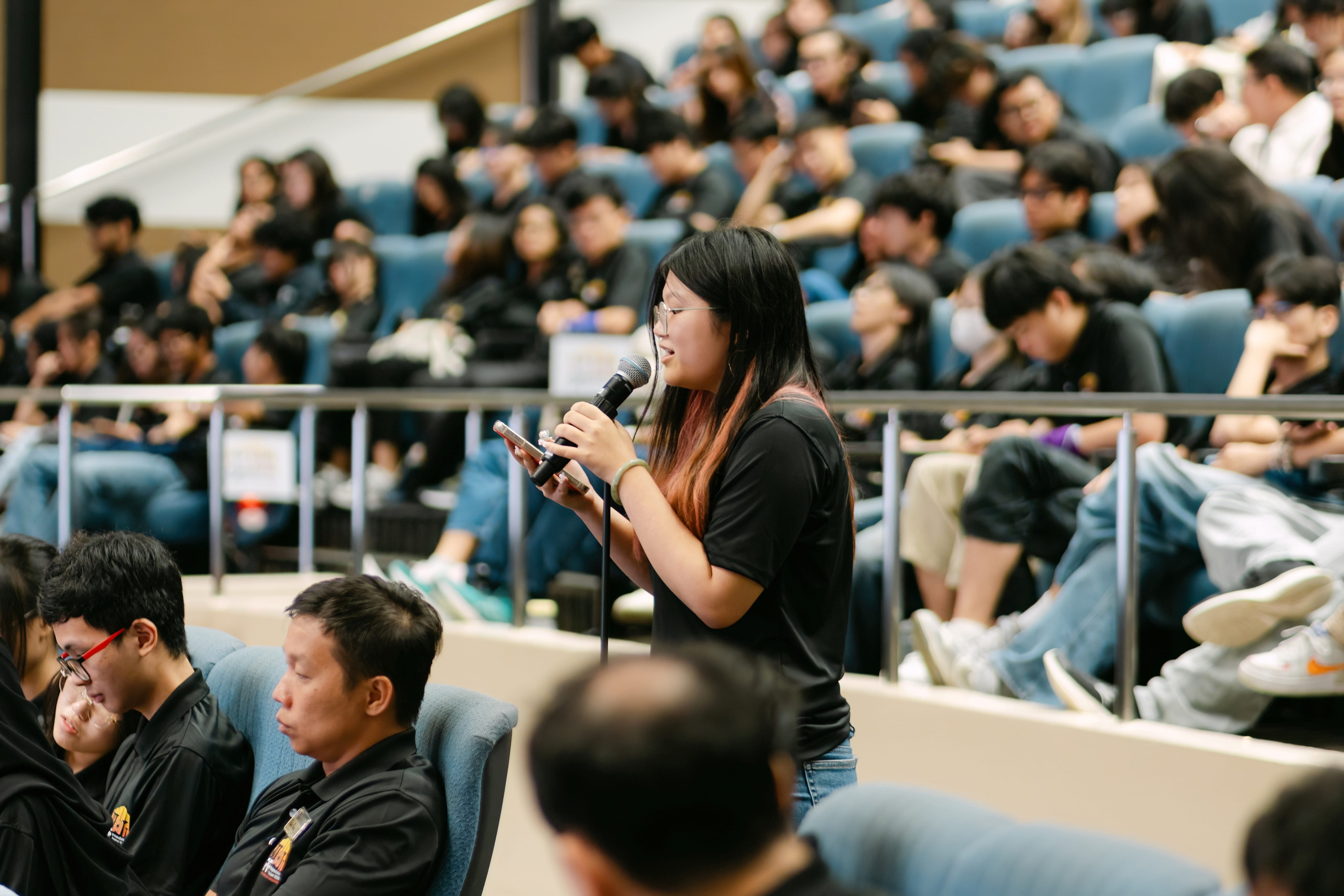 A young woman with glasses speaks into a microphone in a crowded auditorium filled with students in black shirts.
