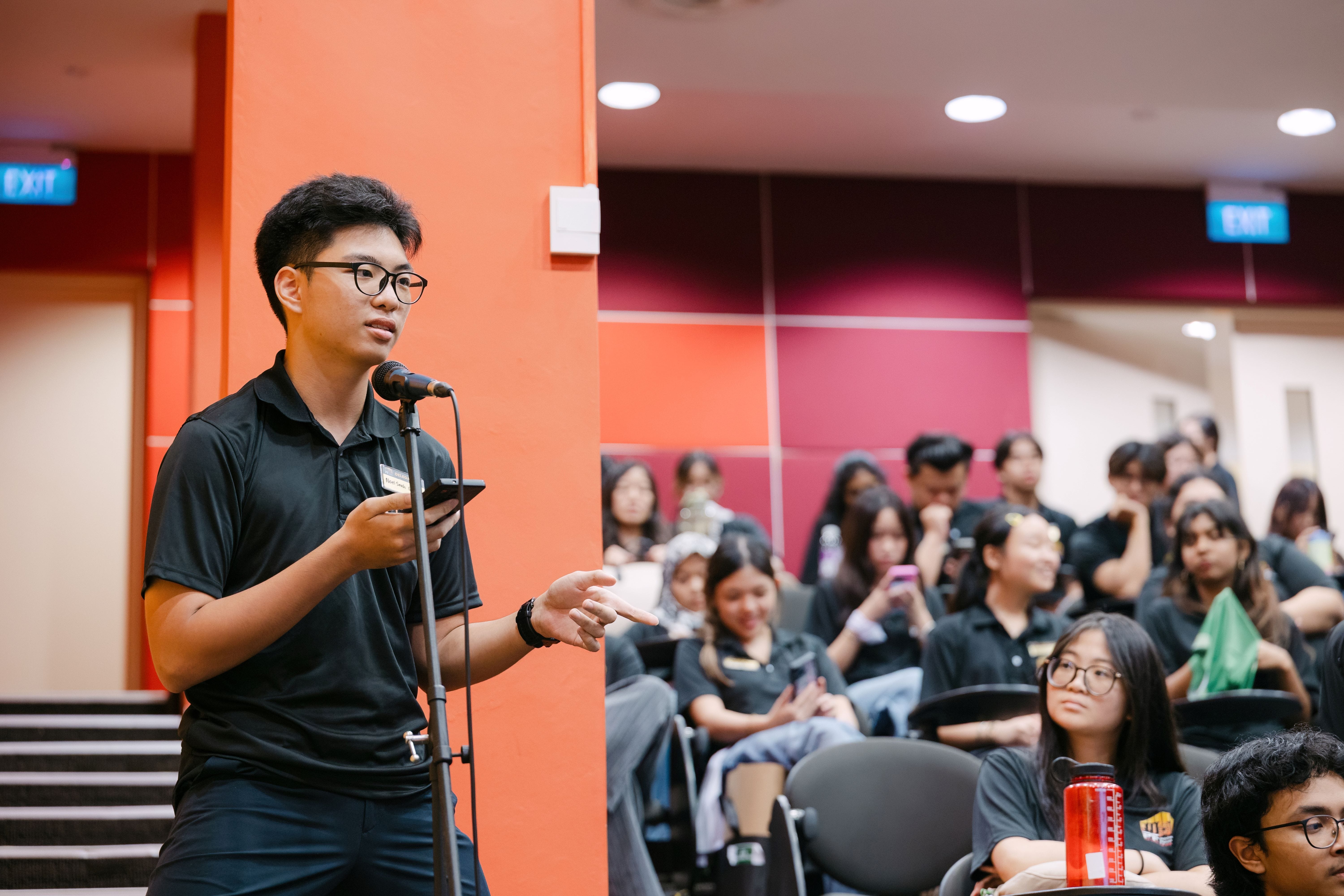 Young man with glasses in a black polo speaks at a microphone stand before a seated audience in black shirts.