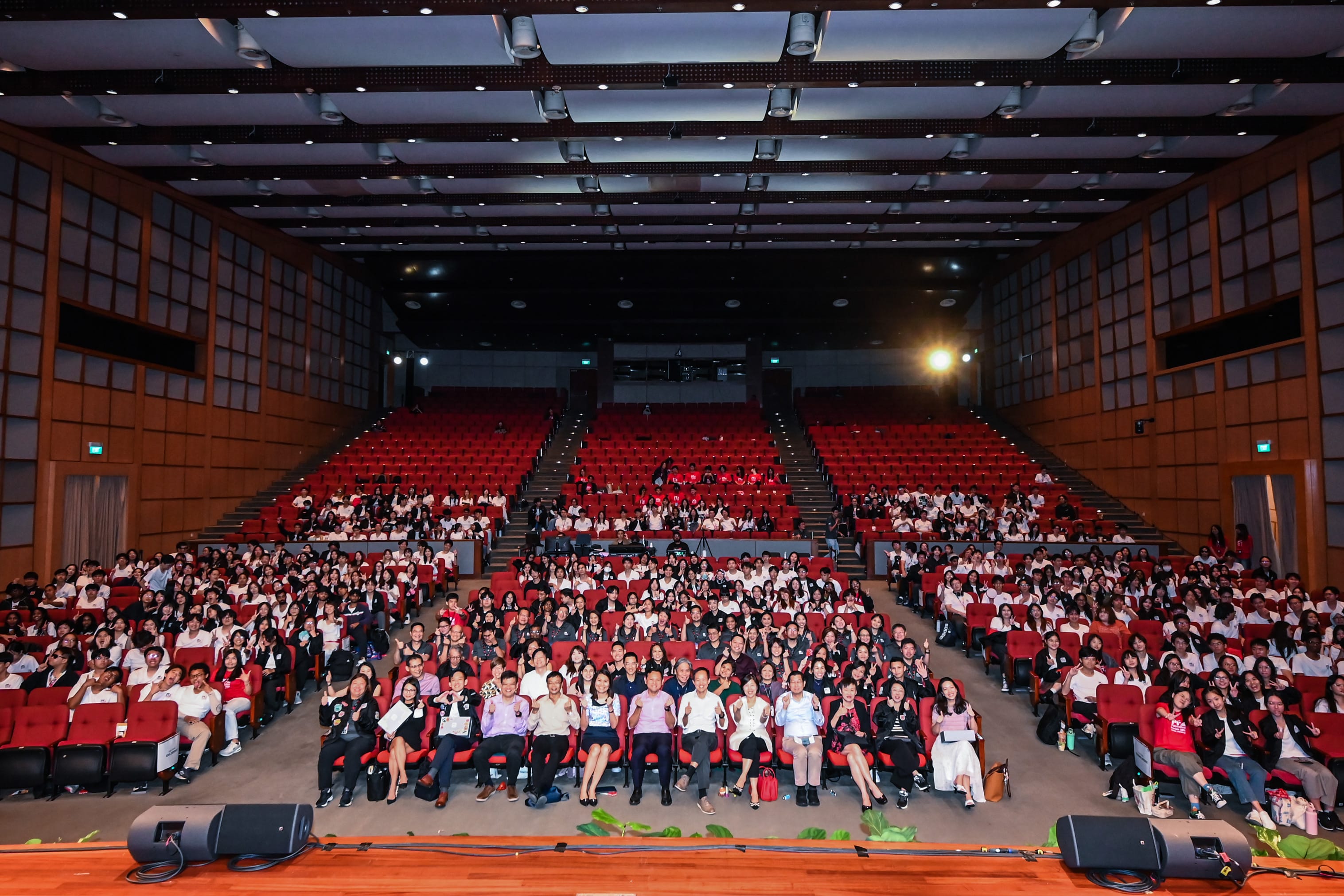 Large auditorium filled with hundreds of people seated in red chairs, posing for a group photo.