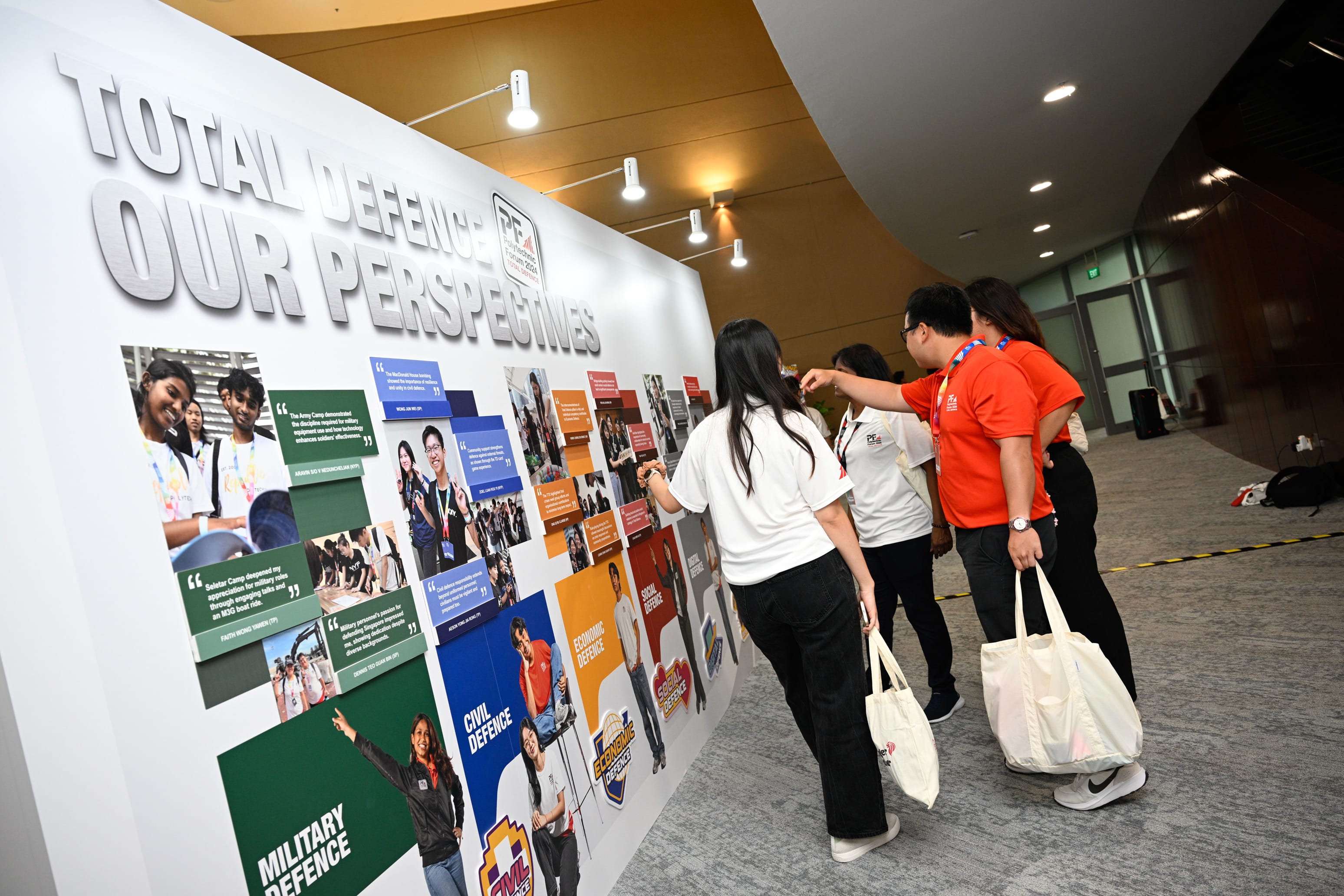 Young people stand before a large display wall reading "Total Defence Our Perspectives" with photos and text panels.