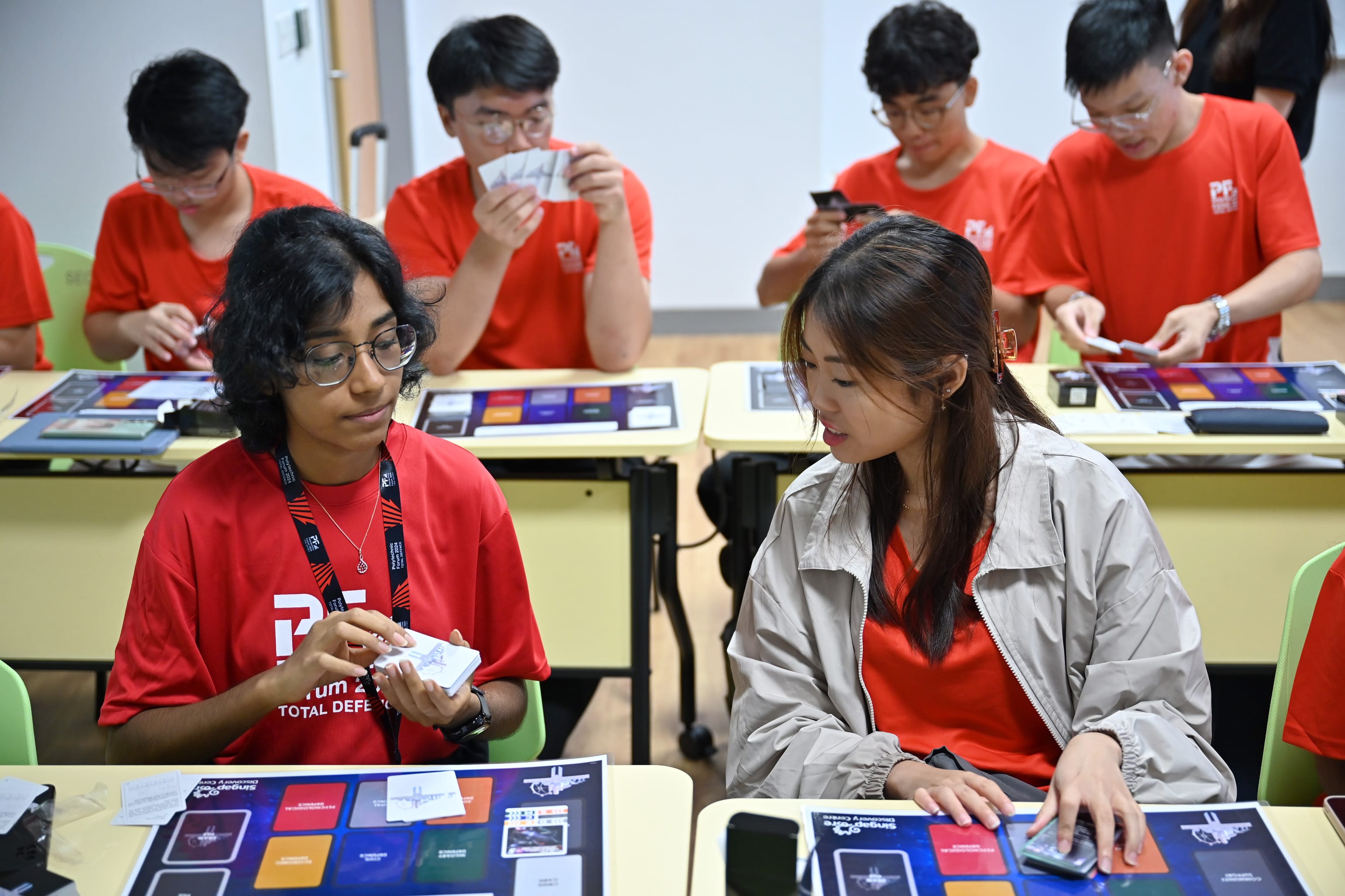 Six young people in red shirts sit at classroom desks playing a card board game with colorful game mats.