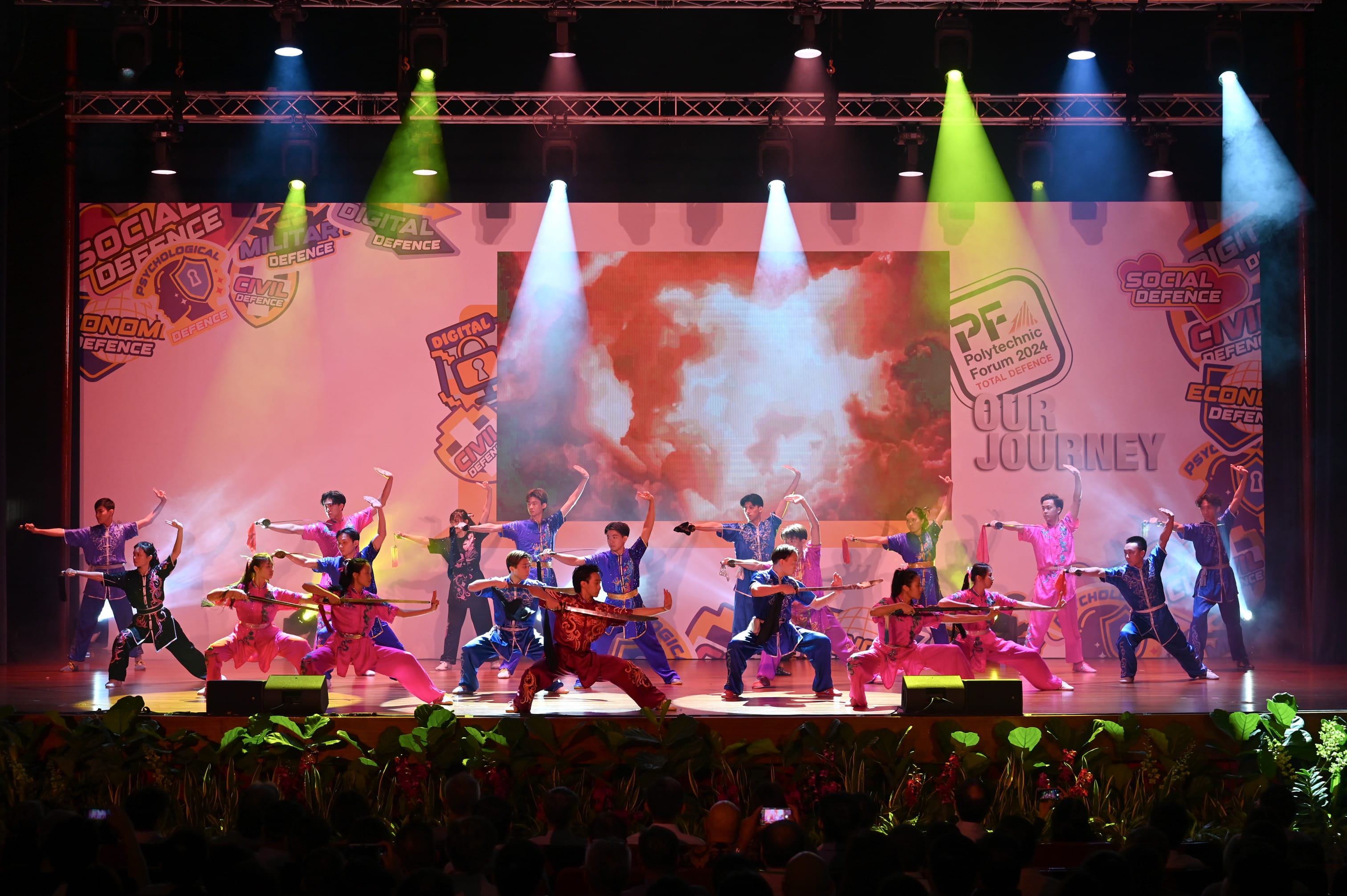 Performers in pink and blue costumes do martial arts poses on a lit stage at Polytechnic Forum 2024.