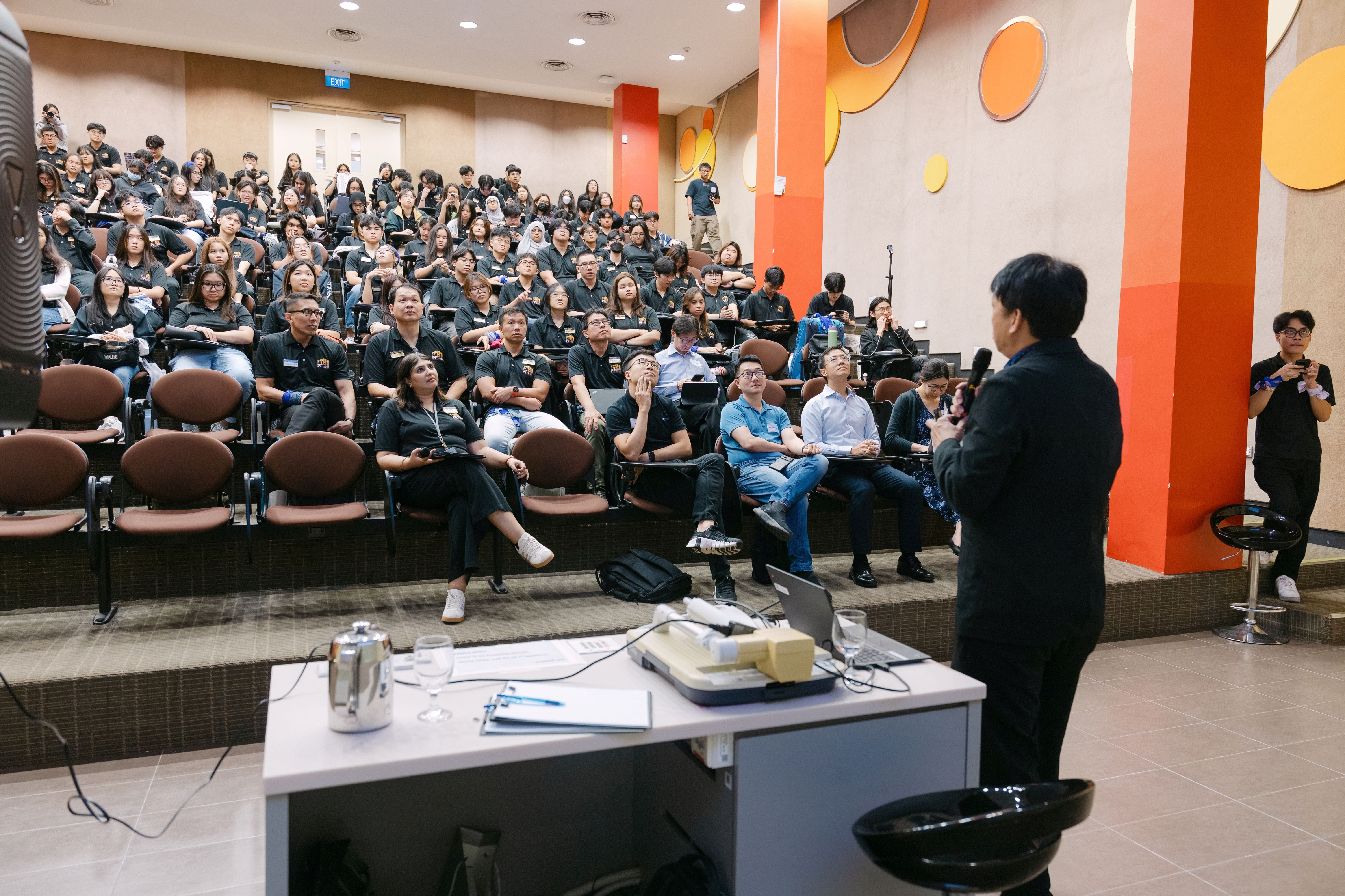 A speaker addresses a large audience in a tiered lecture hall with orange decor and students in black shirts.