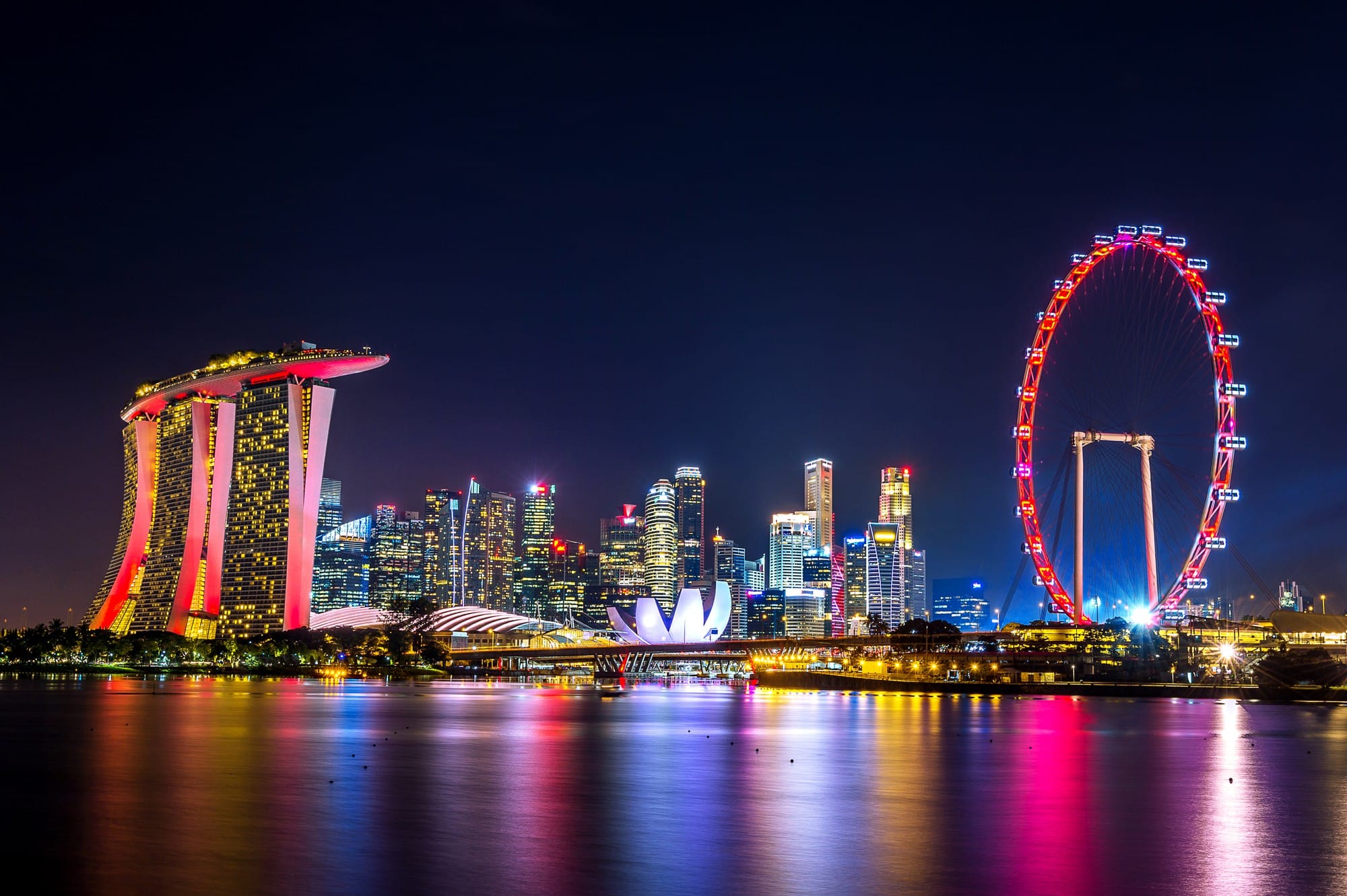 Singapore skyline at night with Marina Bay Sands, a red-lit Ferris wheel, and colorful reflections on the water.