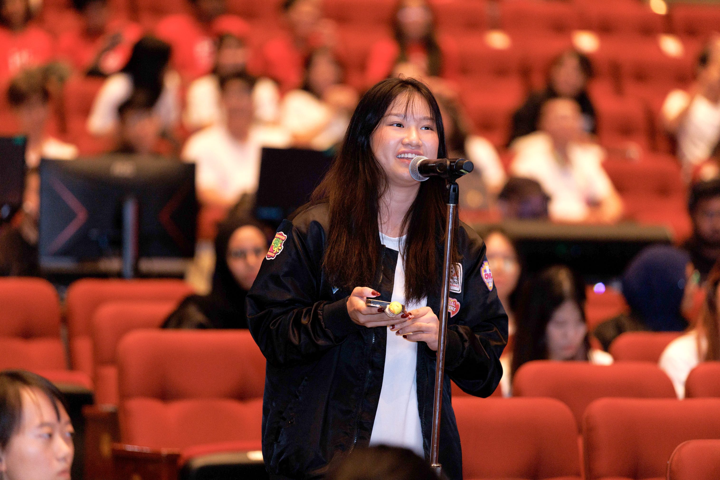 A young woman in a black bomber jacket smiles while speaking into a microphone in a red-seated auditorium.