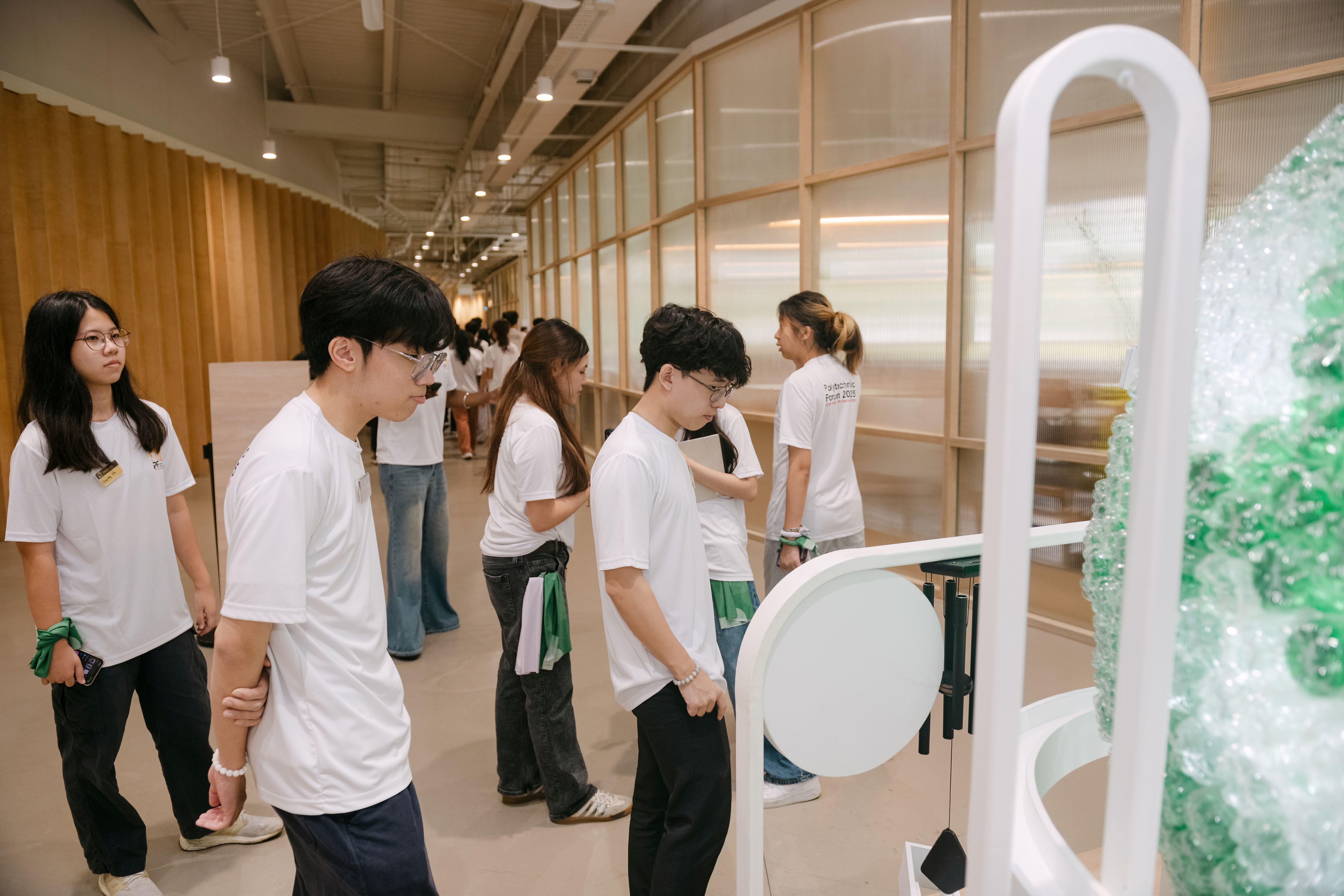Young people in white shirts examine a white sculptural display with green glass pieces in a modern indoor hall.
