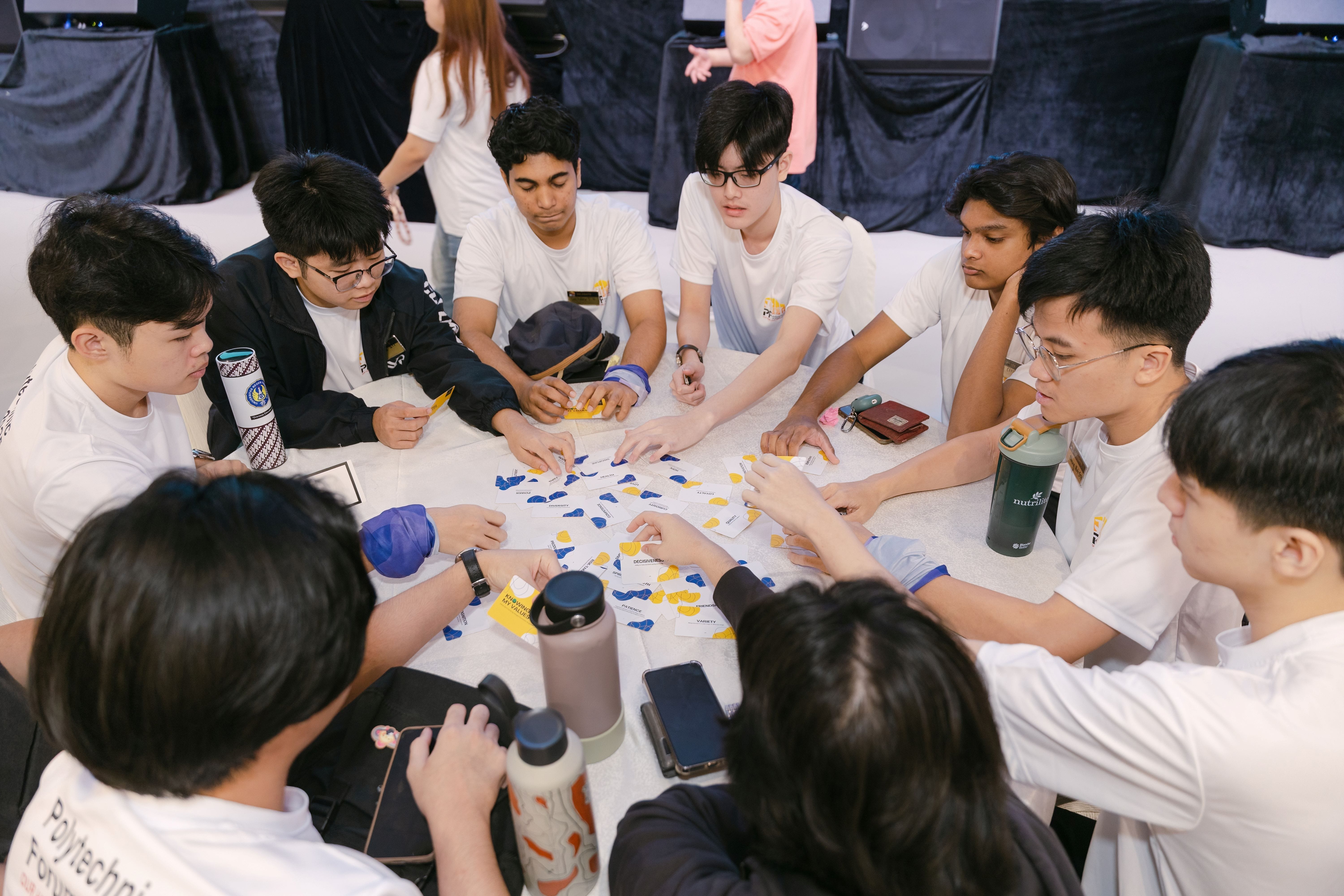 A group of young people in white T-shirts huddle around a table sorting blue and yellow paper cards together.
