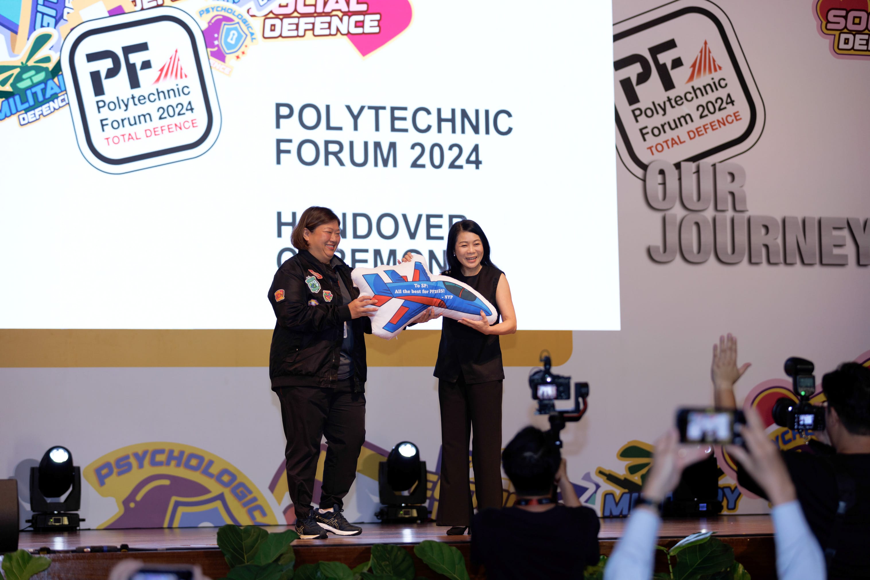 Two women on stage hold a plane-shaped cushion during Polytechnic Forum 2024 Total Defence Handover Ceremony.
