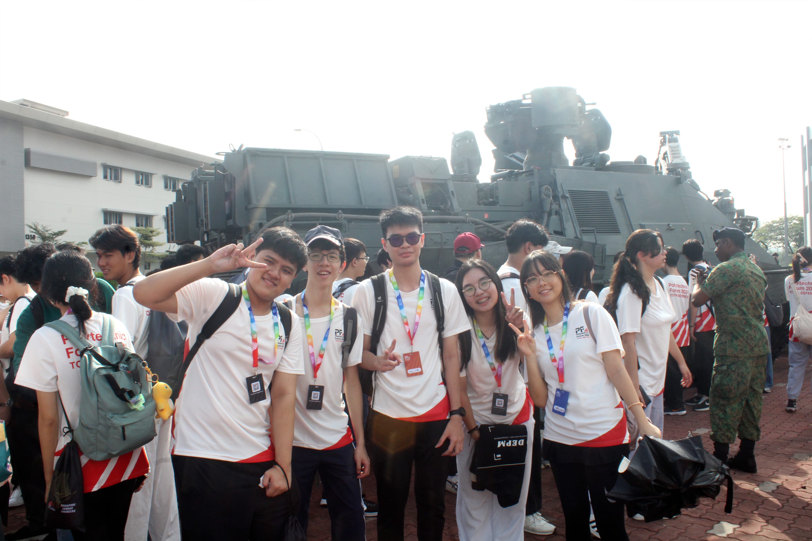 Five students in white shirts with lanyards pose with peace signs in front of a large military armored vehicle.