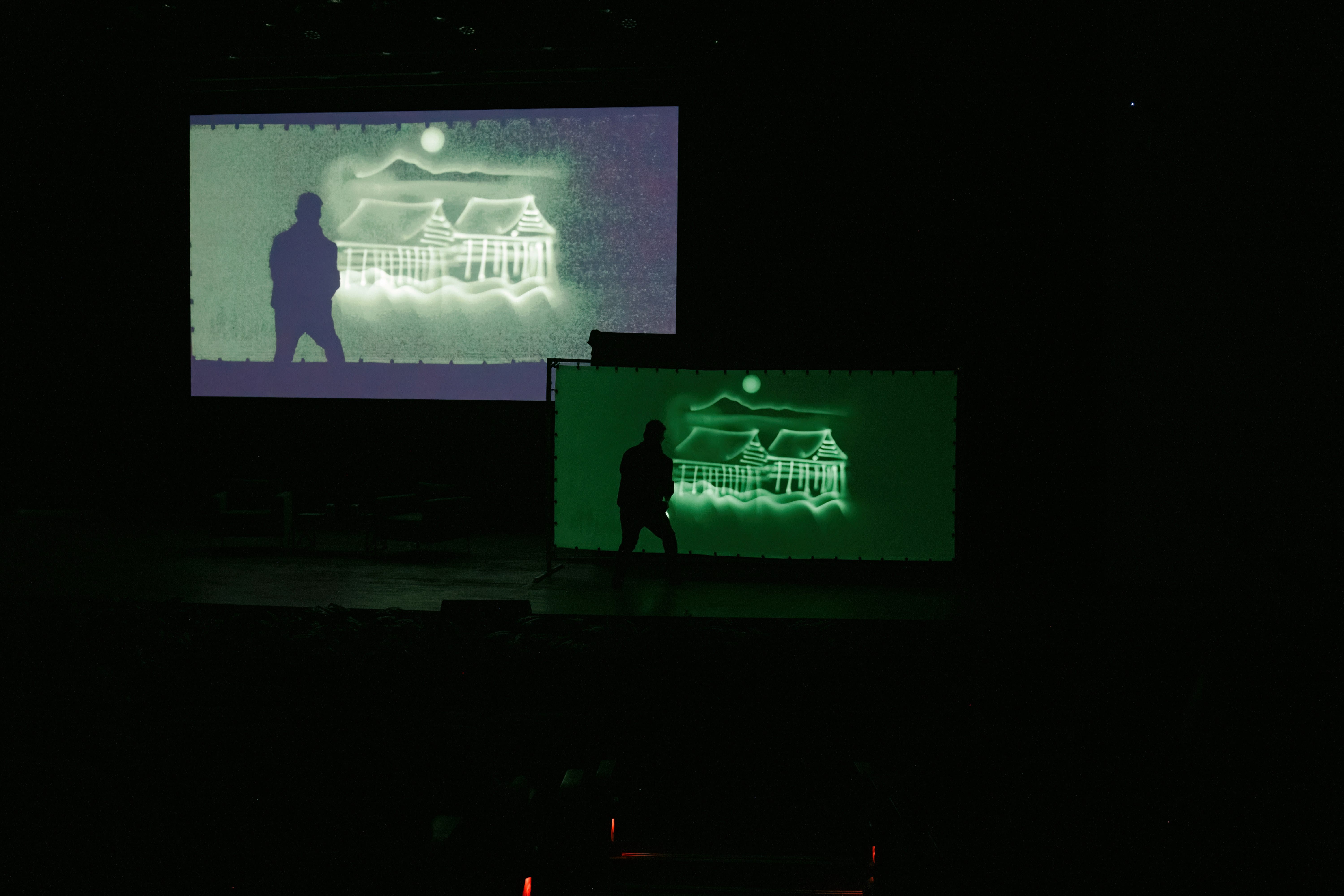 A person's silhouette stands before two green-lit screens showing a building and moon on a dark stage.