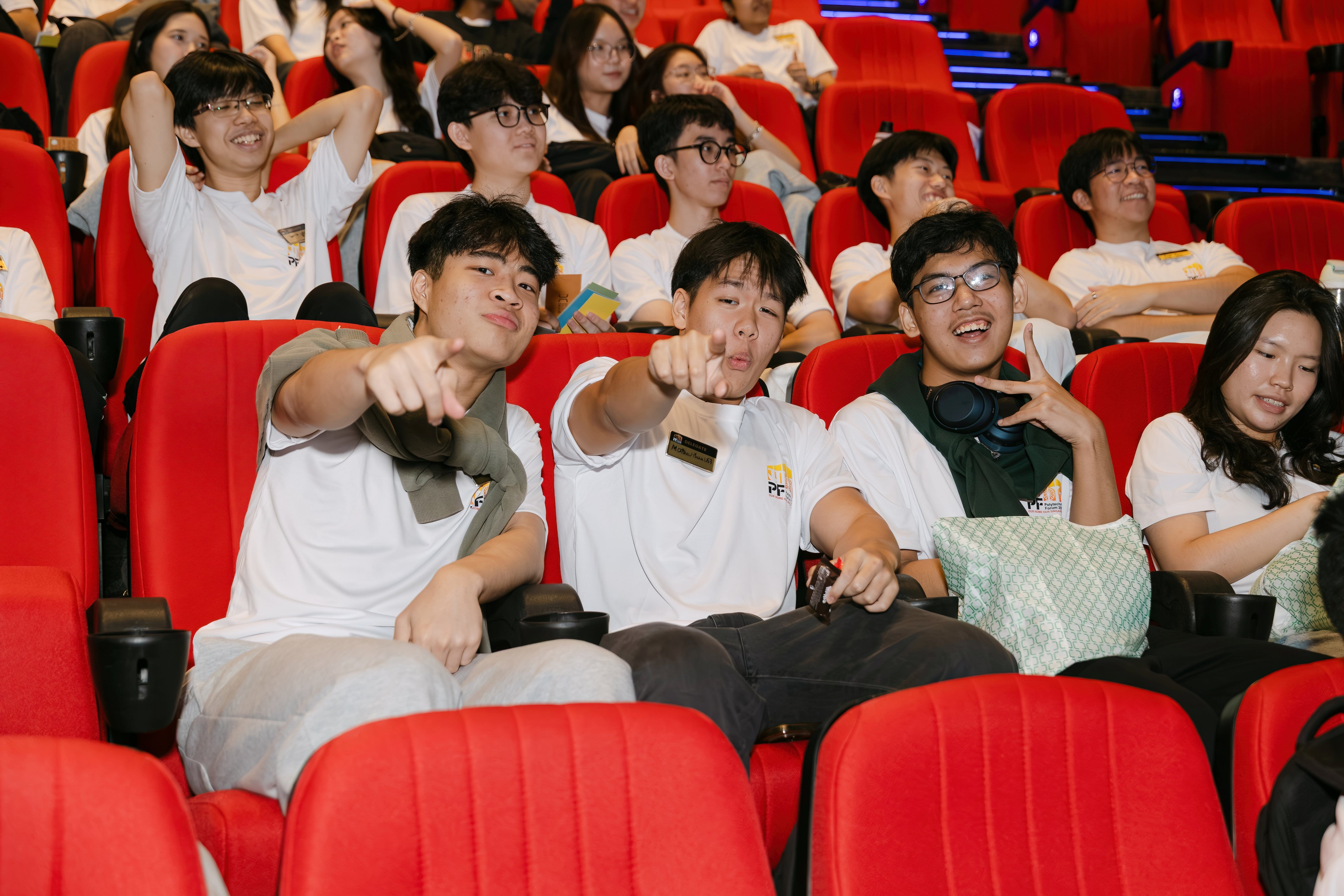 Young people in white T-shirts sit in red theater seats, with two in front pointing and smiling at the camera.