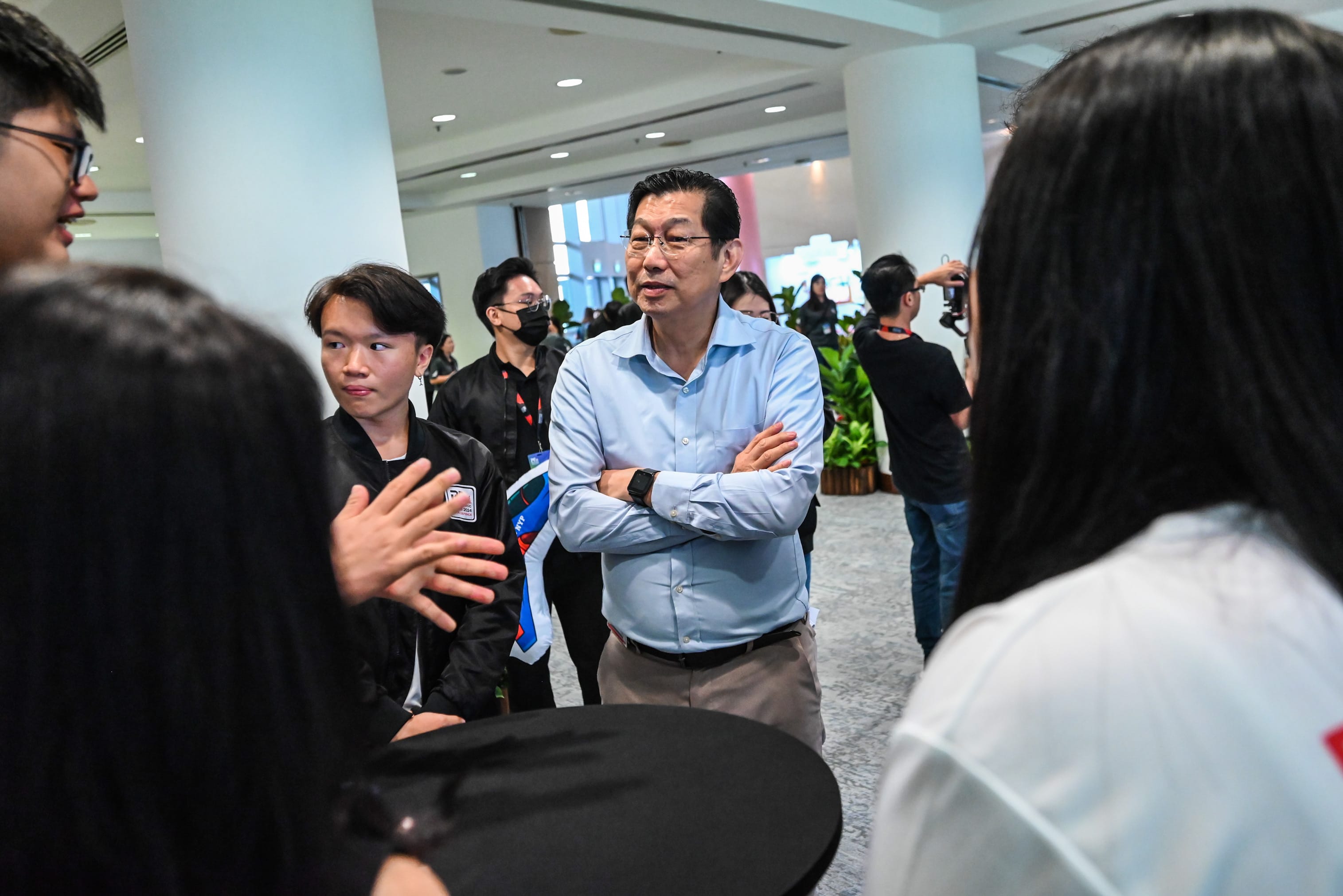 A middle-aged man in a light blue shirt stands with arms crossed, talking with a small group of people indoors.