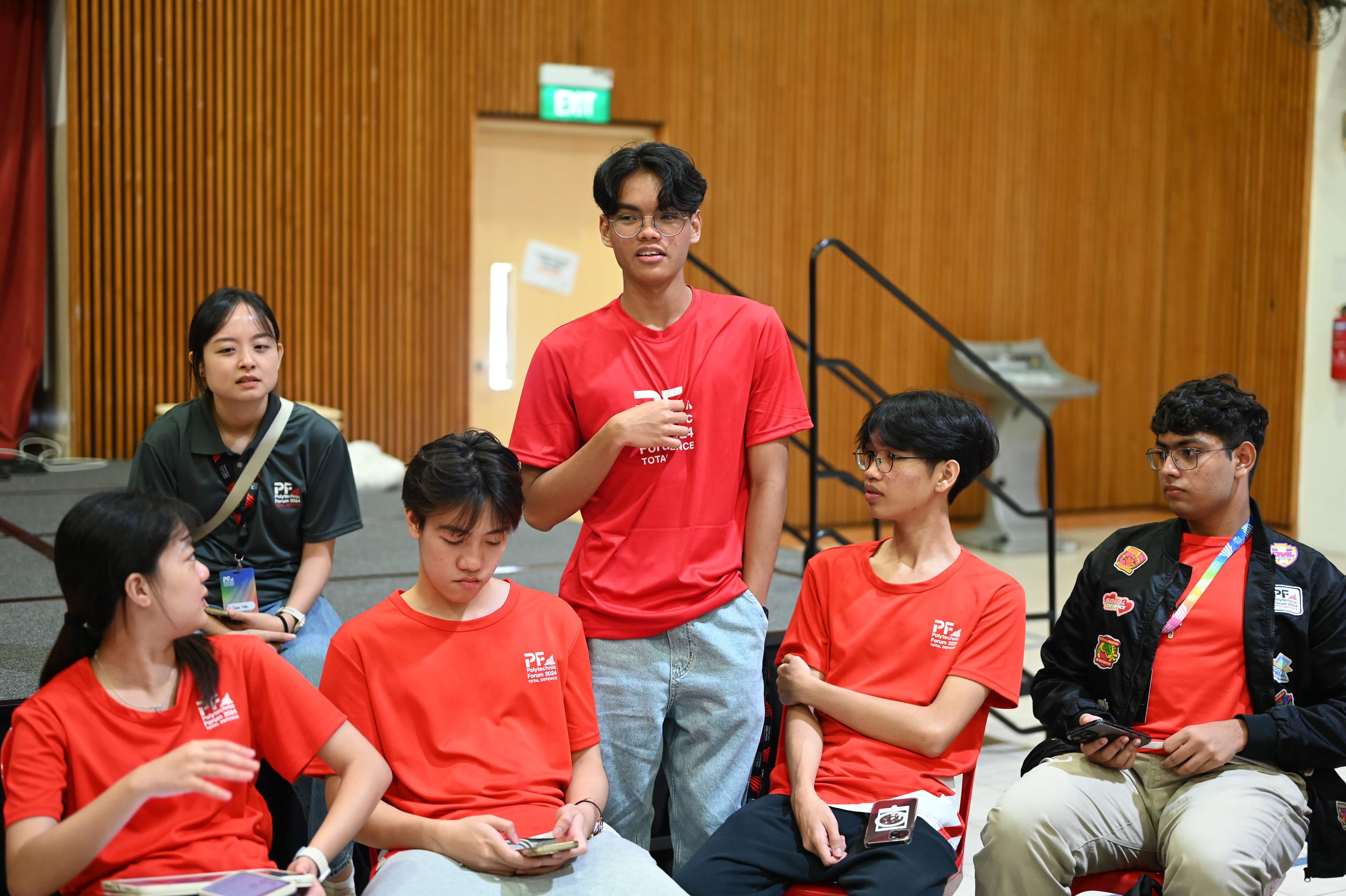 A young man in a red PF shirt stands speaking to a seated group of students in matching red shirts indoors.