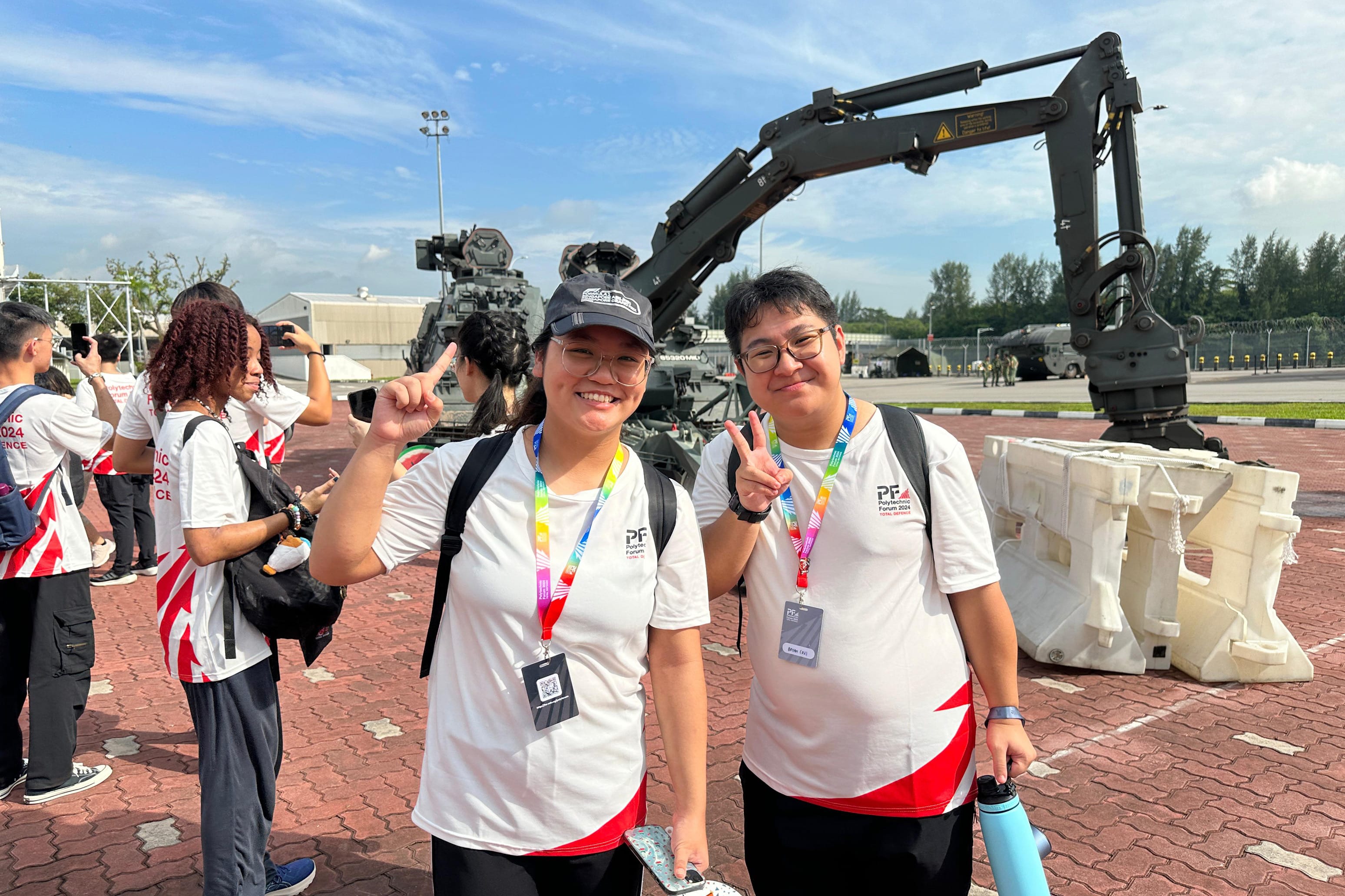 Two smiling students in white Polytechnic Forum 2024 shirts pose with hand gestures in front of a large military robotic arm.