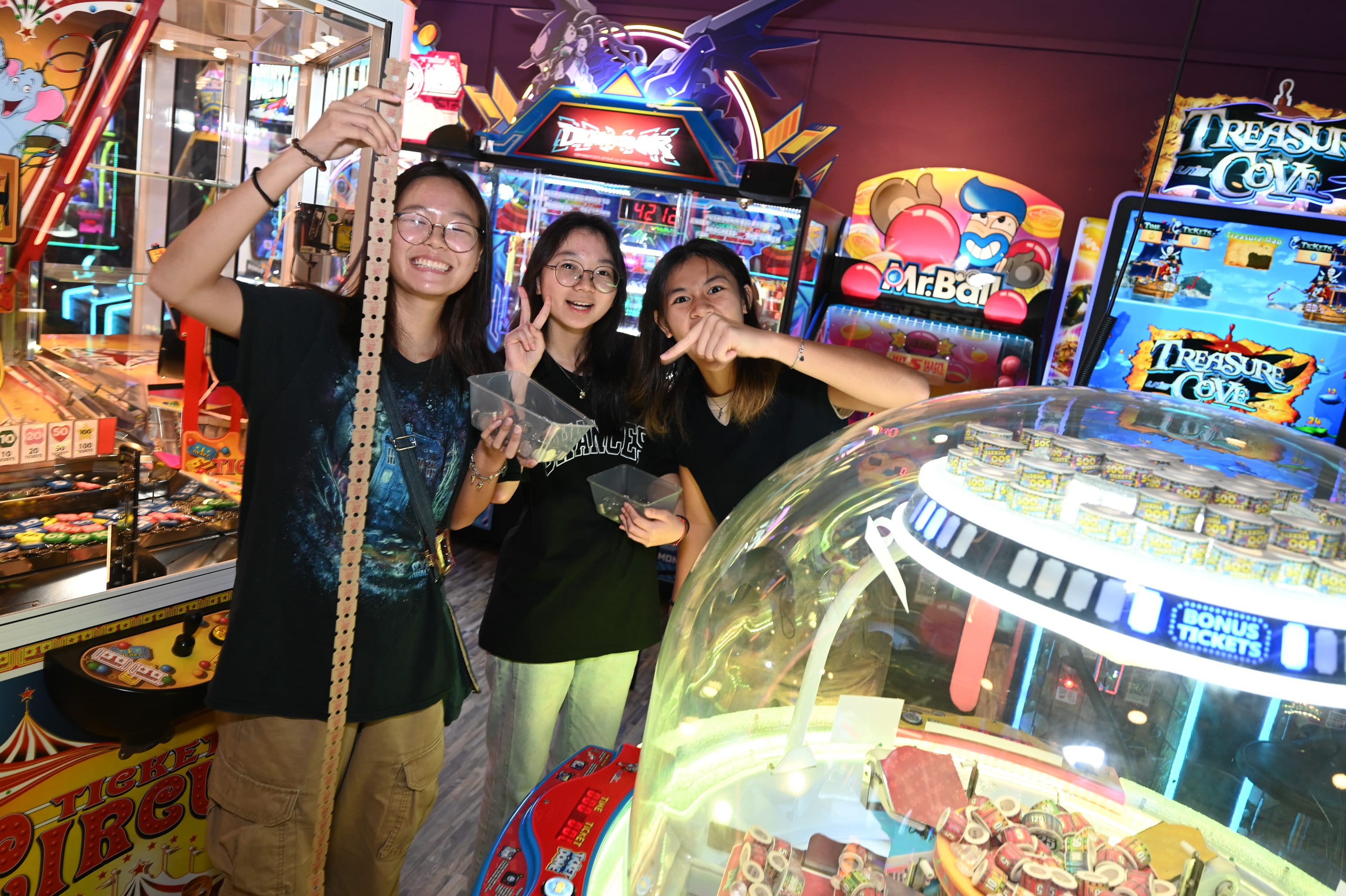 Three smiling young women pose in a colorful arcade, one holding a long strip of tickets near glowing game machines.