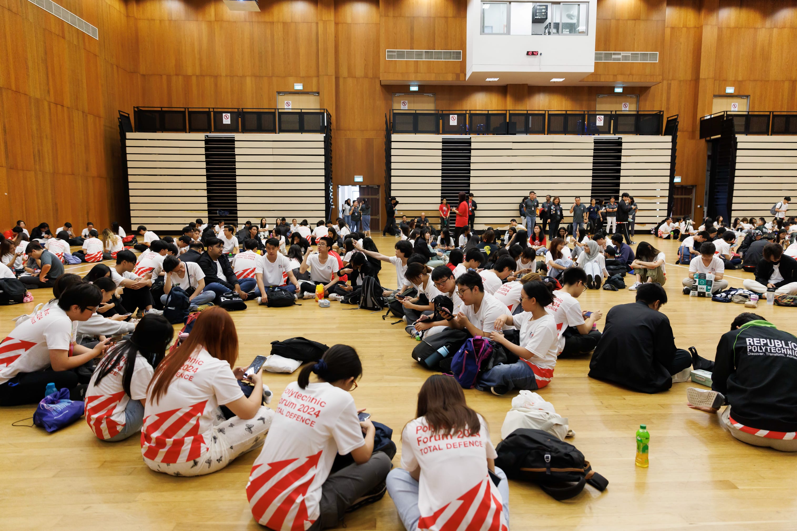 Large group of students in Polytechnic Forum 2024 Total Defence shirts sitting on a wooden gym floor.