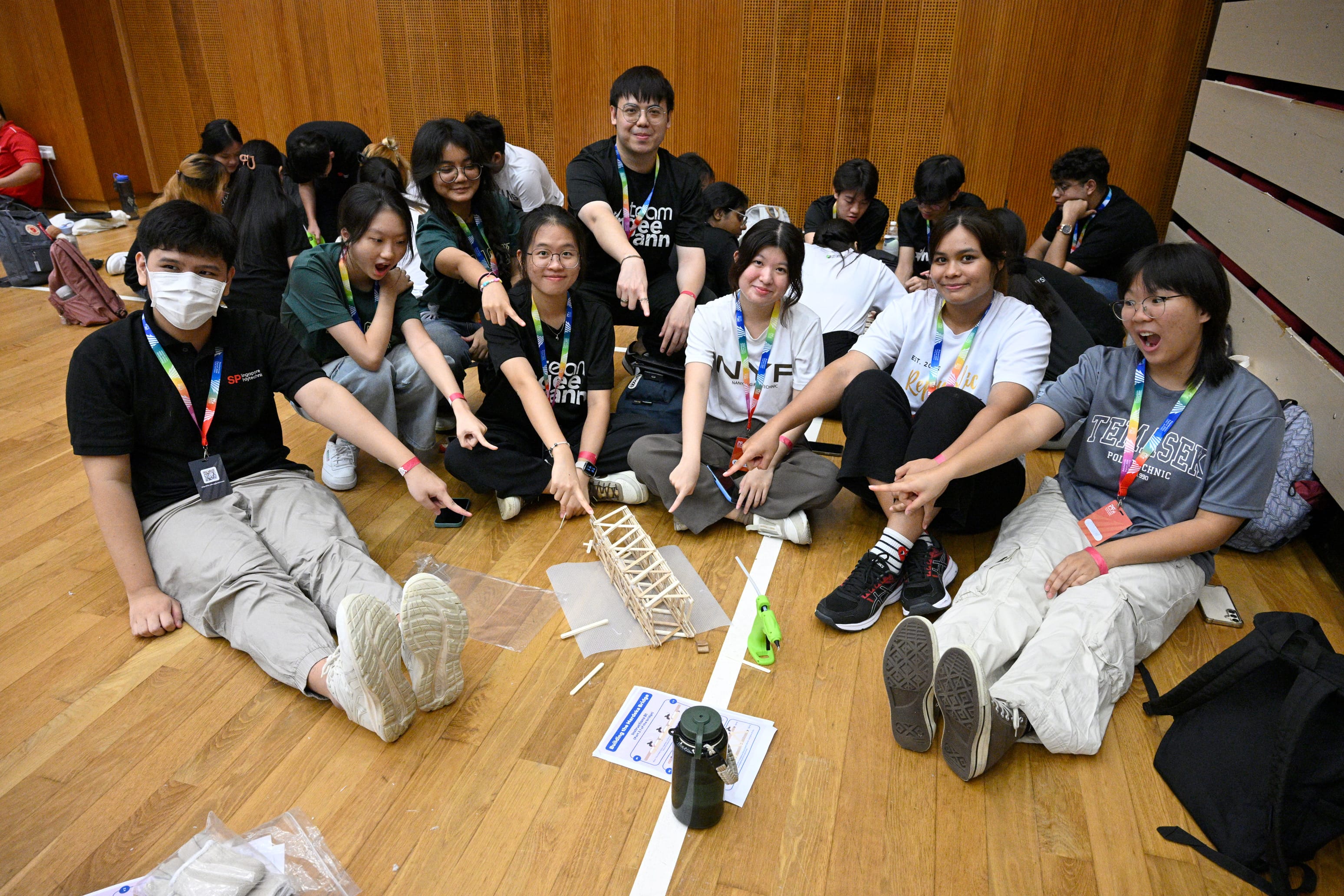 A group of students sitting on a wooden floor, pointing at a small handmade popsicle stick bridge they built together.