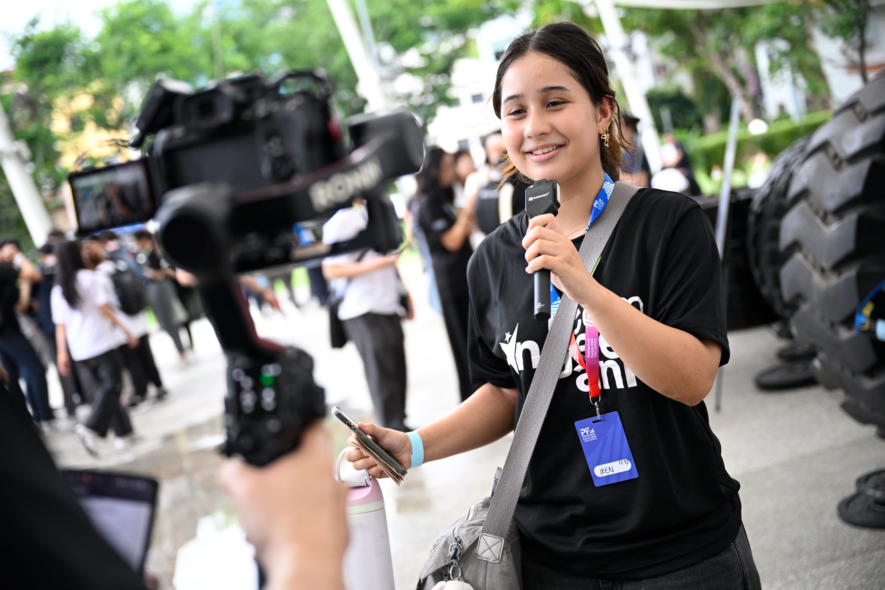 Young woman in a black shirt holds a microphone and smiles at a camera during an outdoor event.