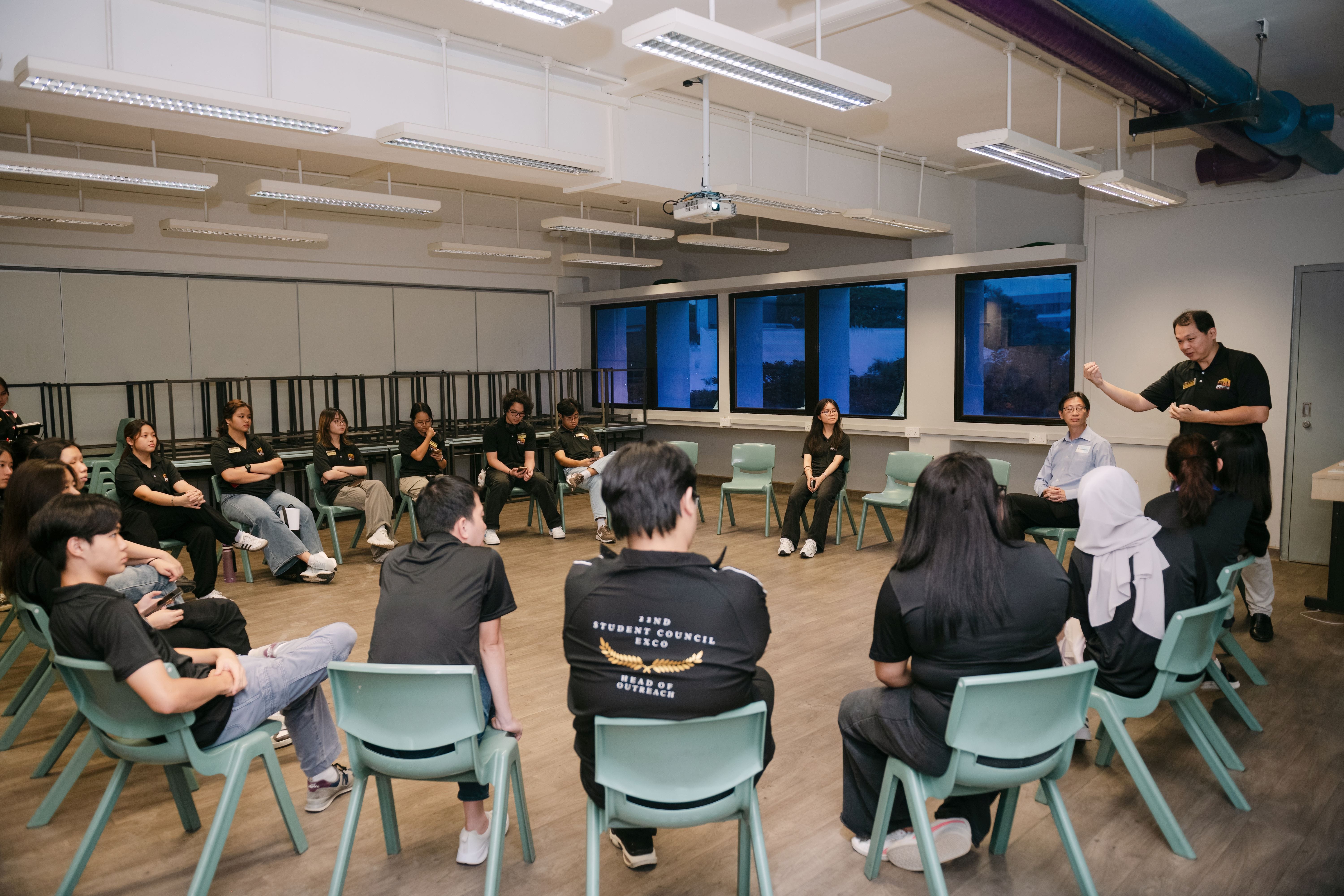 Students in black shirts sit in a circle in a classroom while a standing man addresses the group.