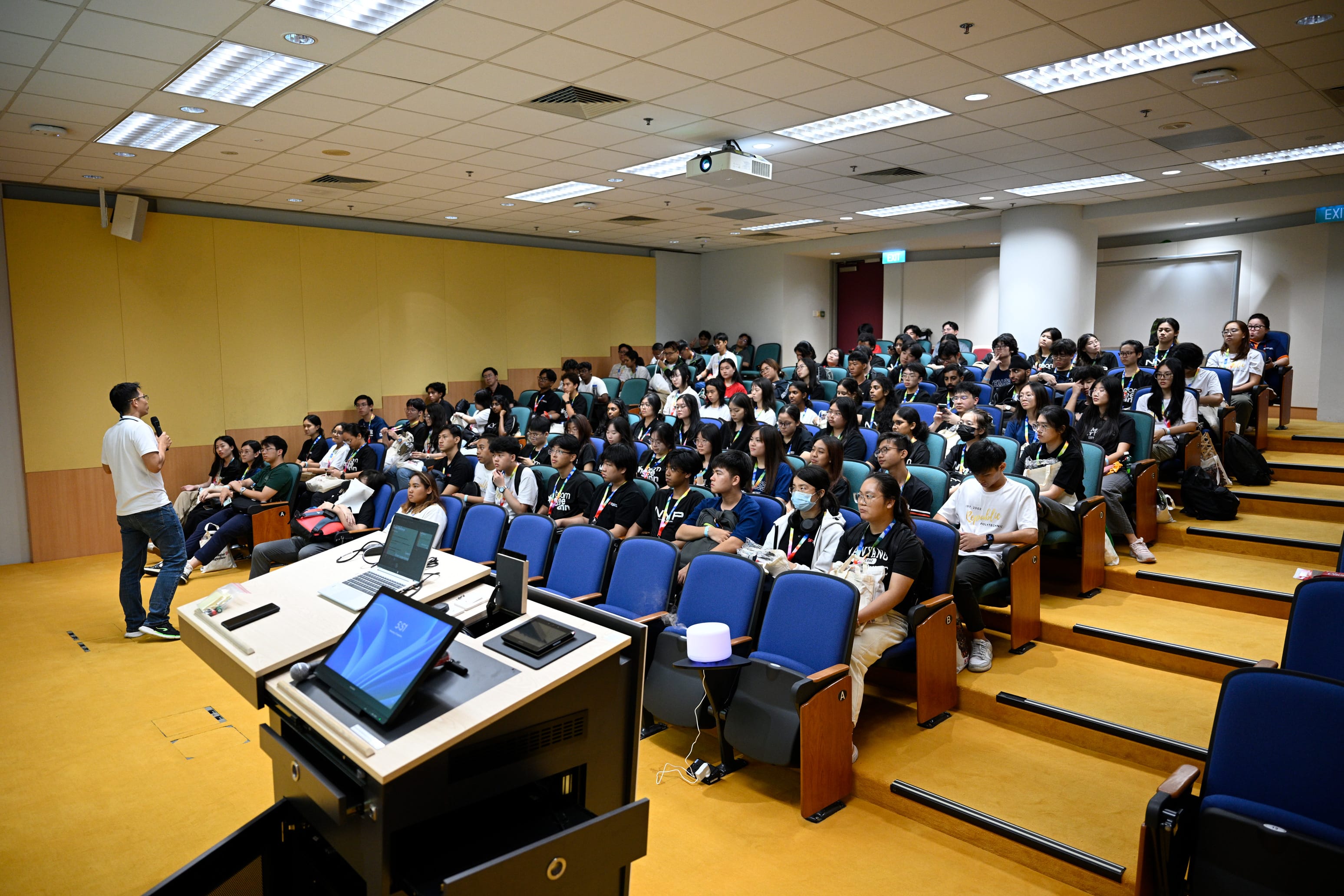 A speaker with a microphone stands before a large audience of students seated in a lecture hall with blue chairs.