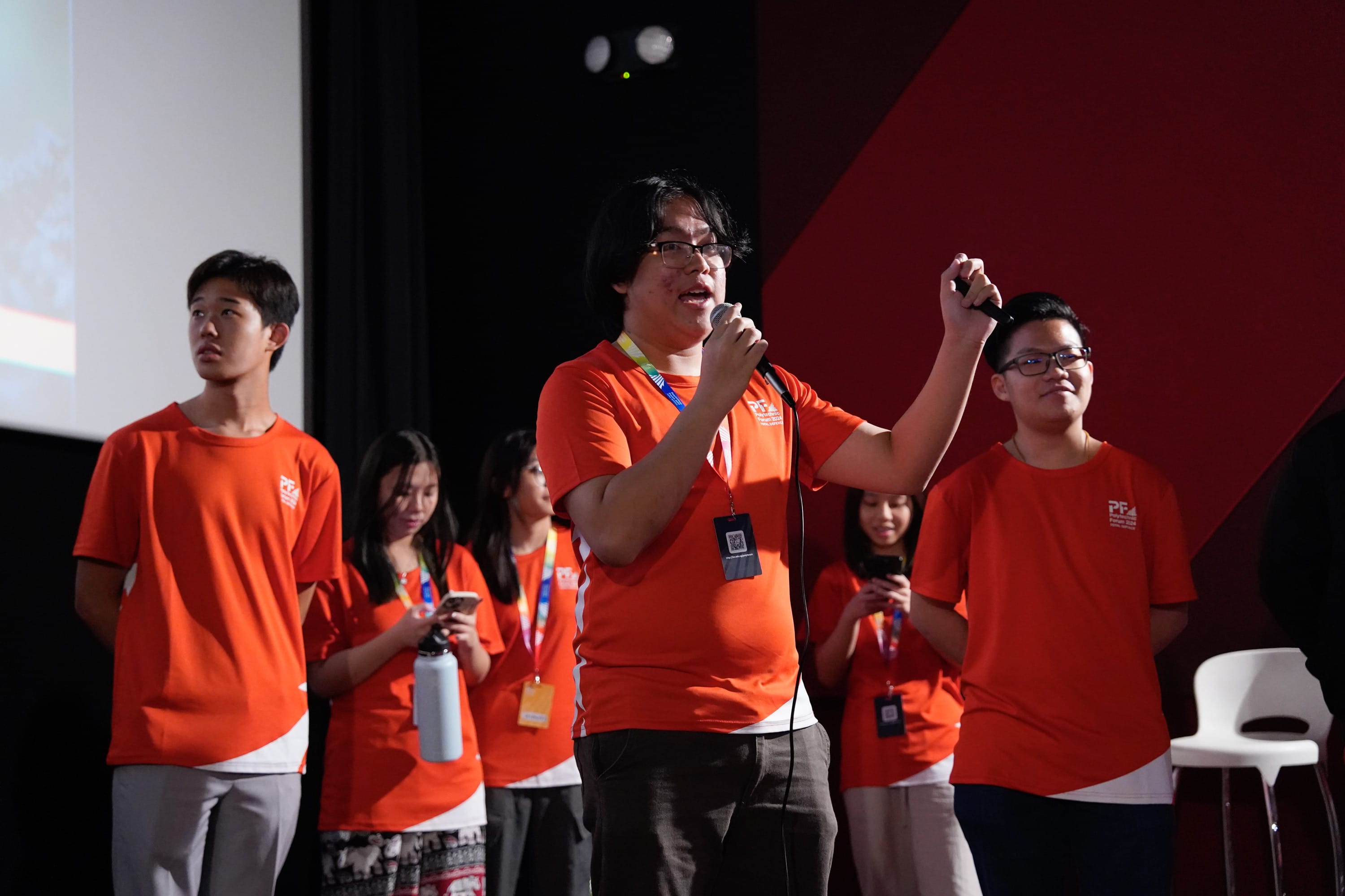 A young man in an orange shirt speaks into a microphone on stage, surrounded by five others in matching orange shirts.