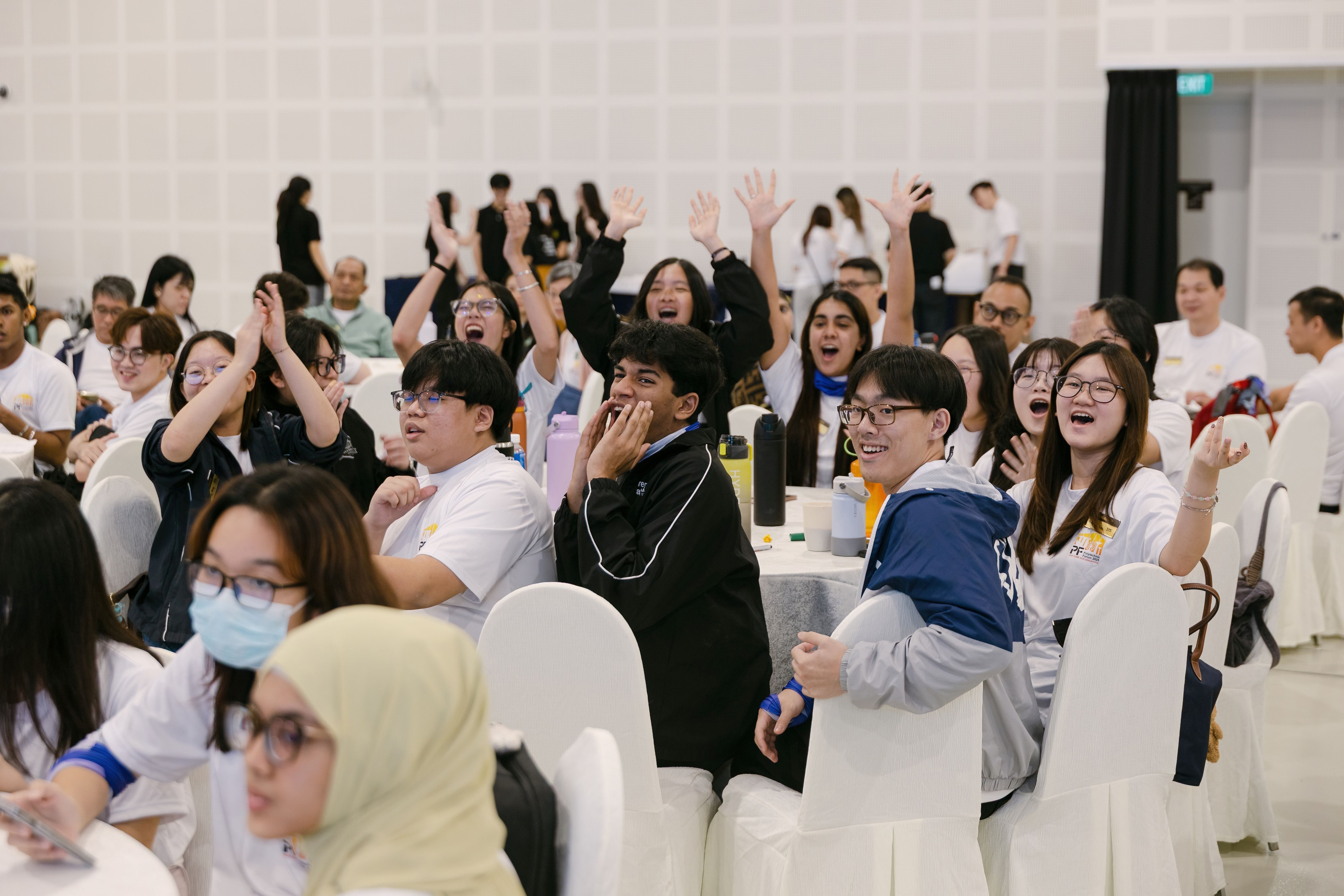 A large group of young people seated at round tables cheering excitedly with hands raised in a bright hall.