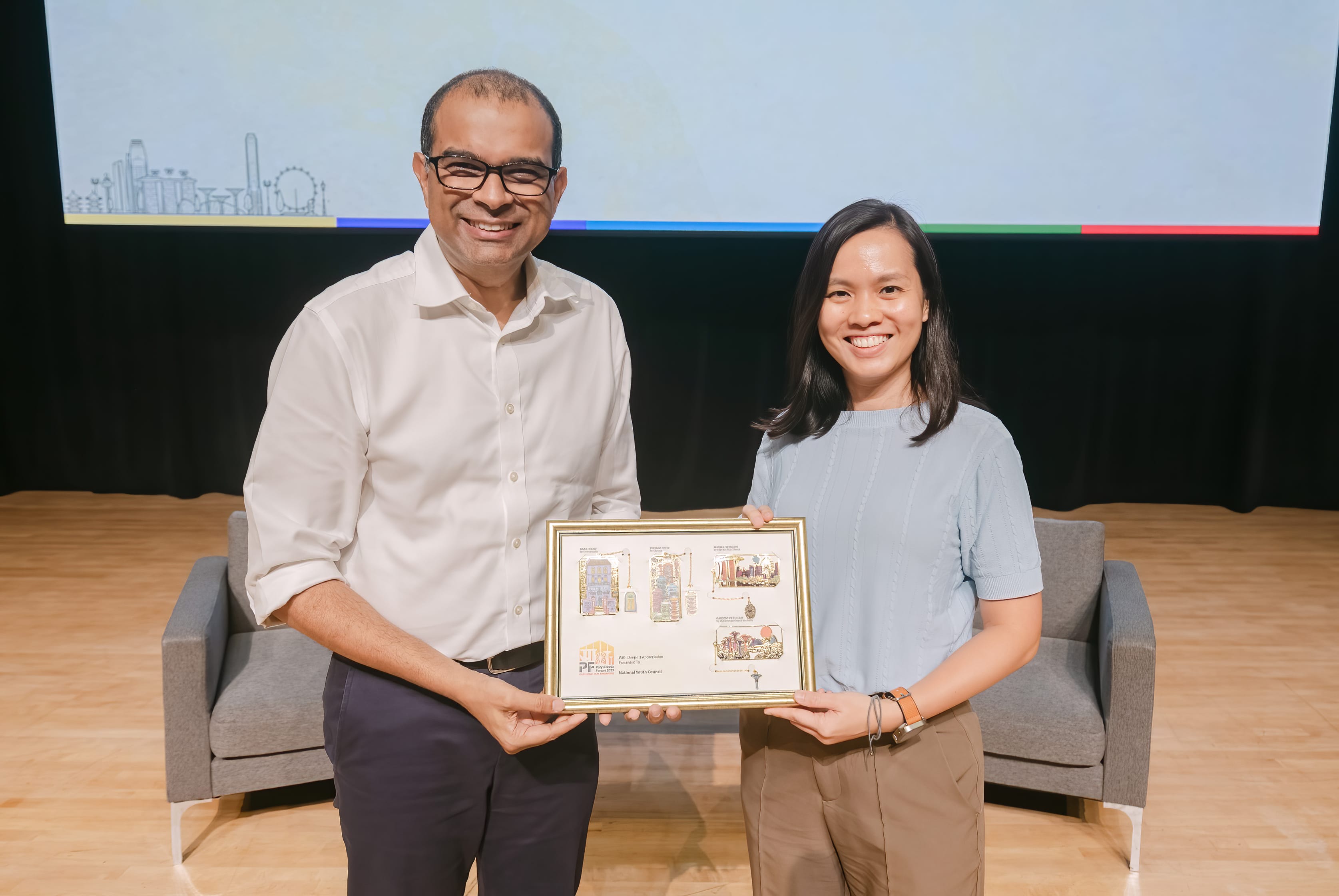 Two smiling people hold a gold-framed plaque together on a stage with a presentation screen behind them.