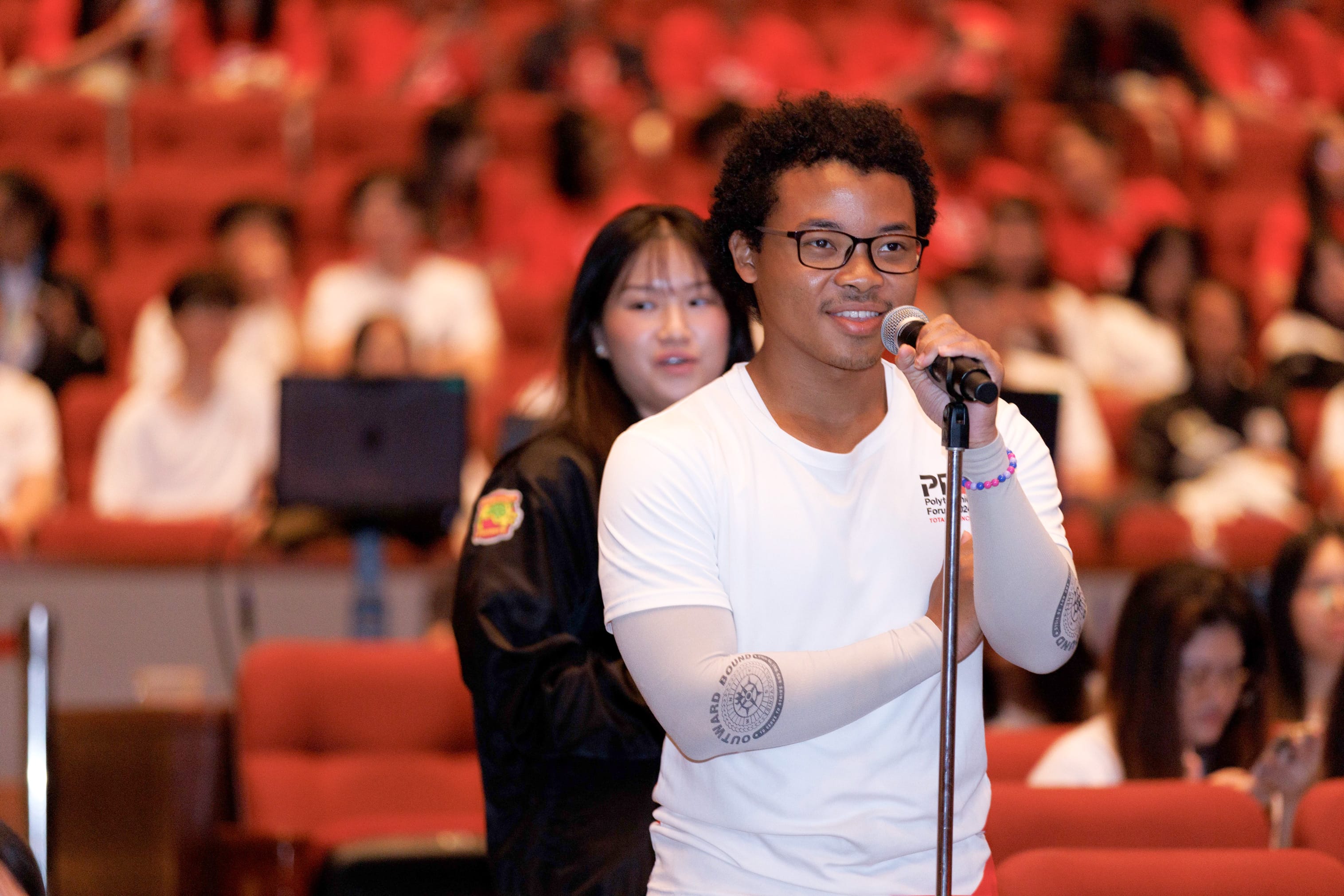 Young man with glasses and curly hair speaks into a microphone in a large auditorium with red seats.