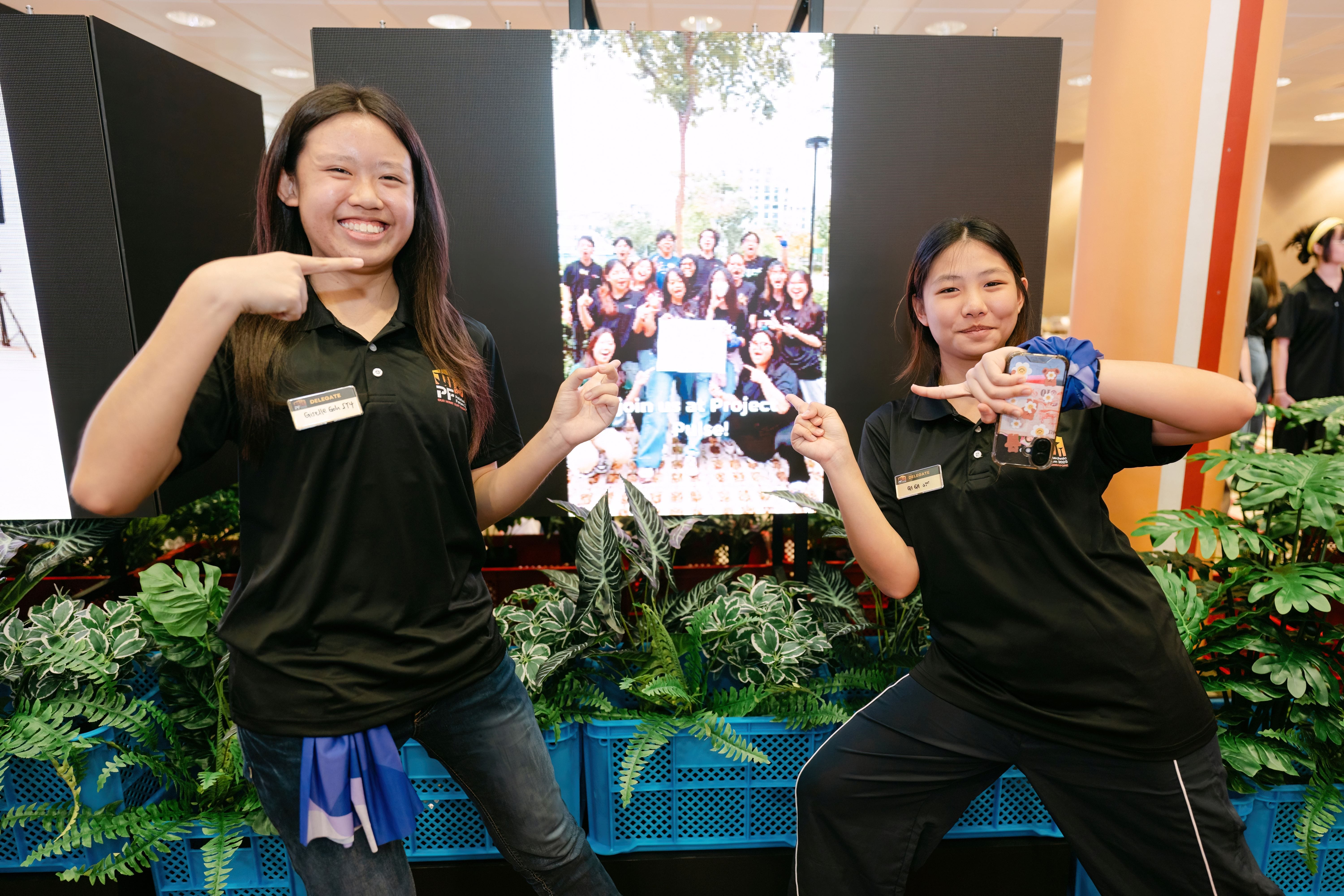Two young women in black polo shirts pose smiling, pointing at each other, in front of a group photo display.