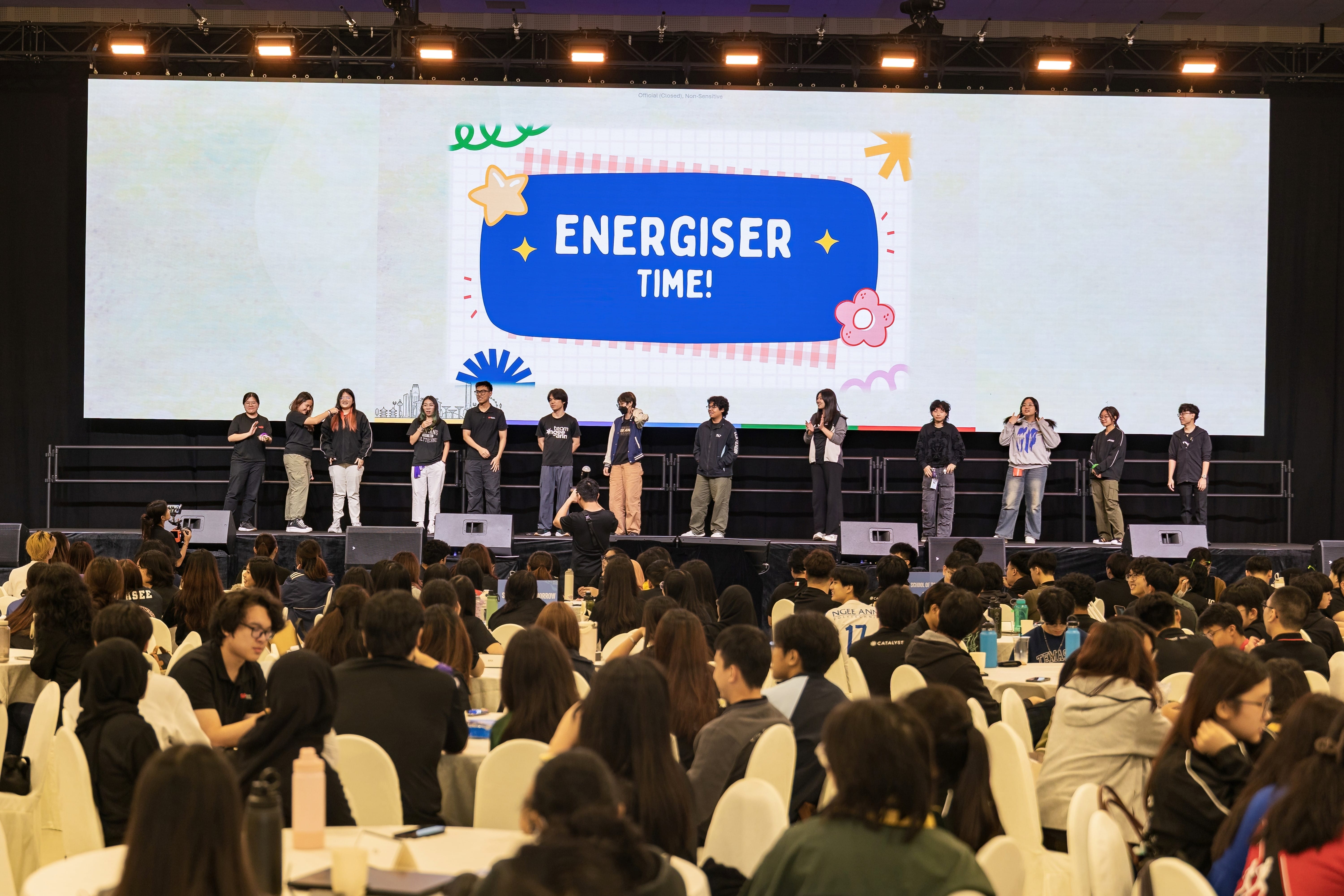 A large indoor event with people on stage and a screen displaying "Energiser Time!" to a seated crowd.