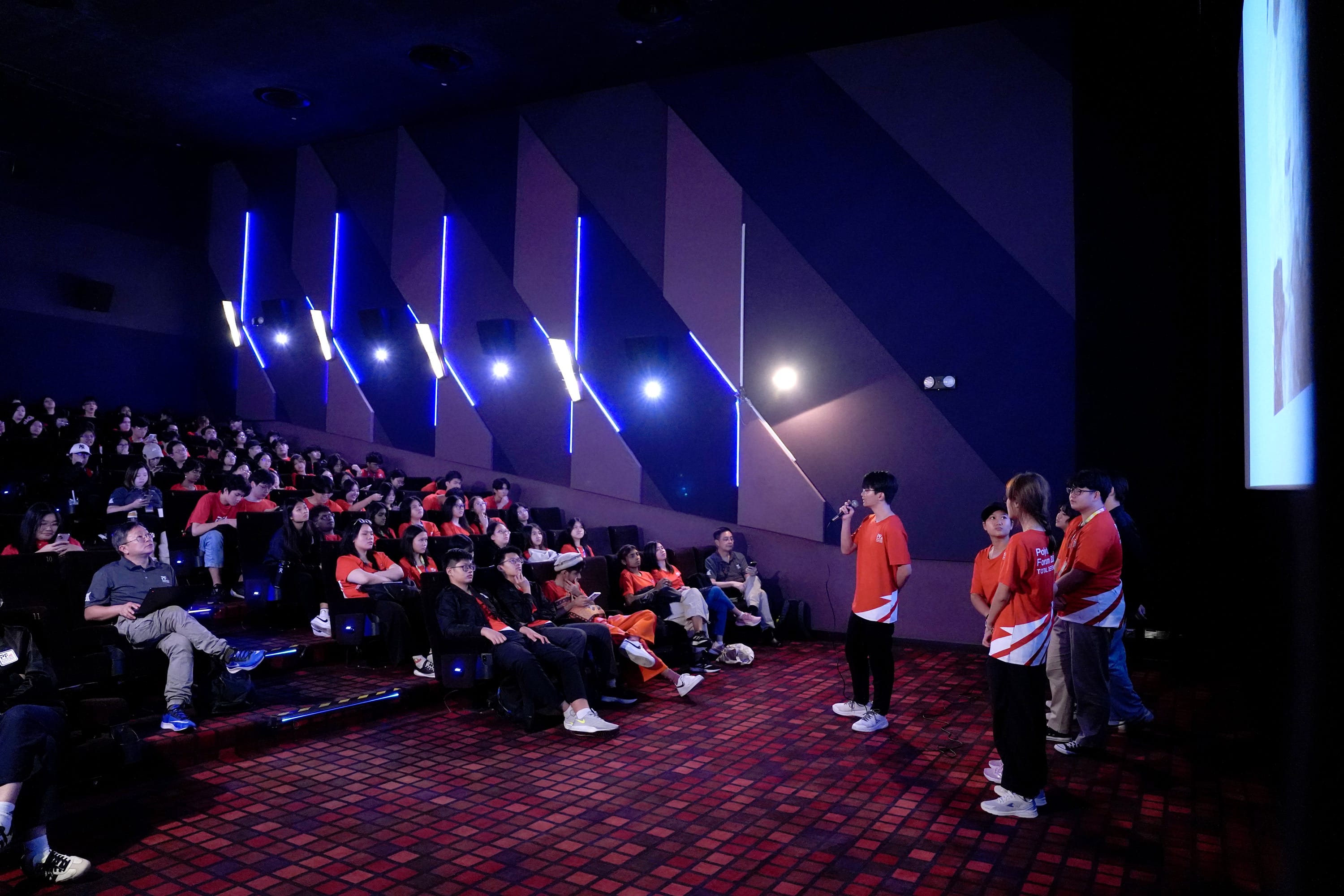 A person in a red shirt speaks with a microphone before a large seated audience in a dark auditorium with blue LED lights.