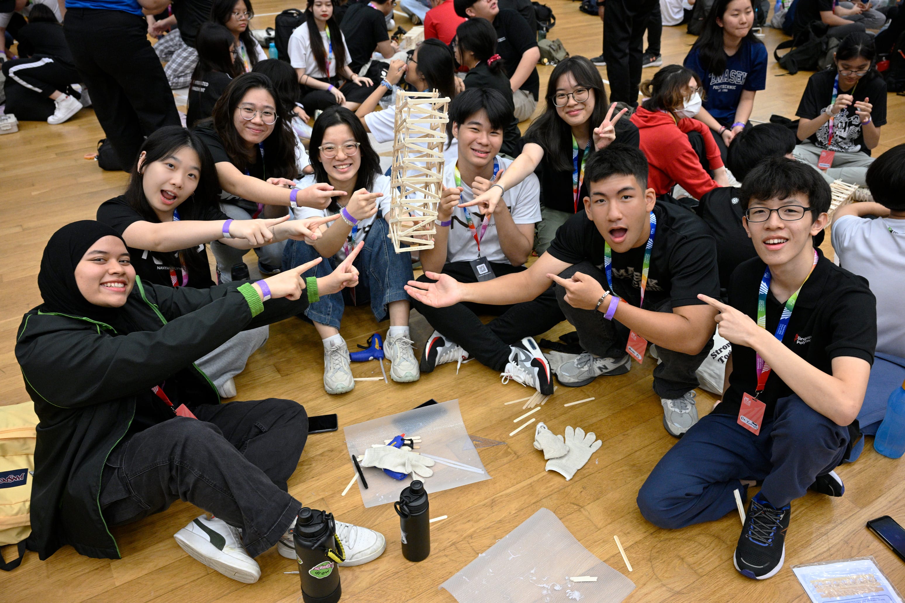 A group of smiling students sit on a gym floor pointing at a small wooden stick tower they built together.