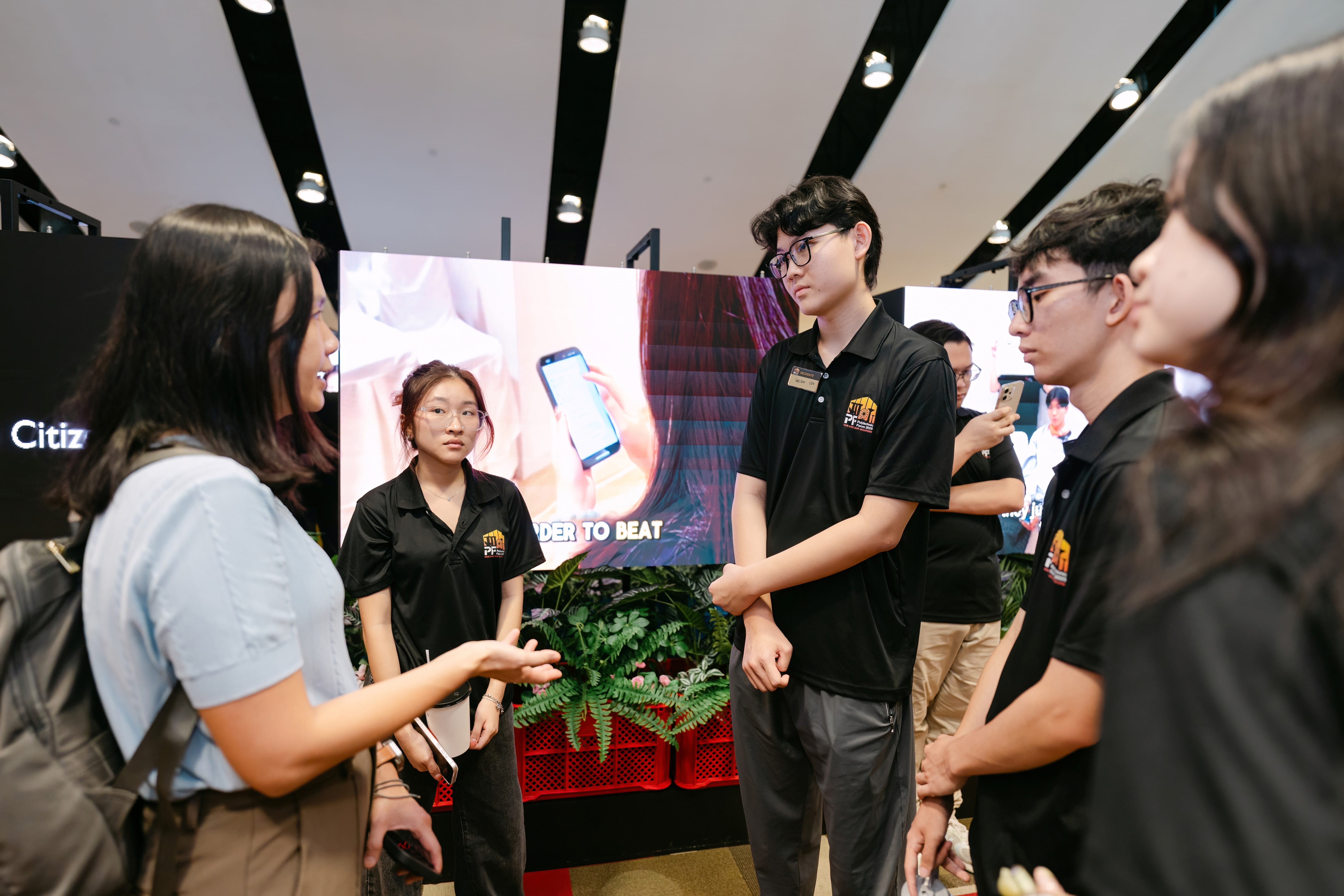 Several young people in black polo shirts talk with a woman in a blue top at an indoor event booth.