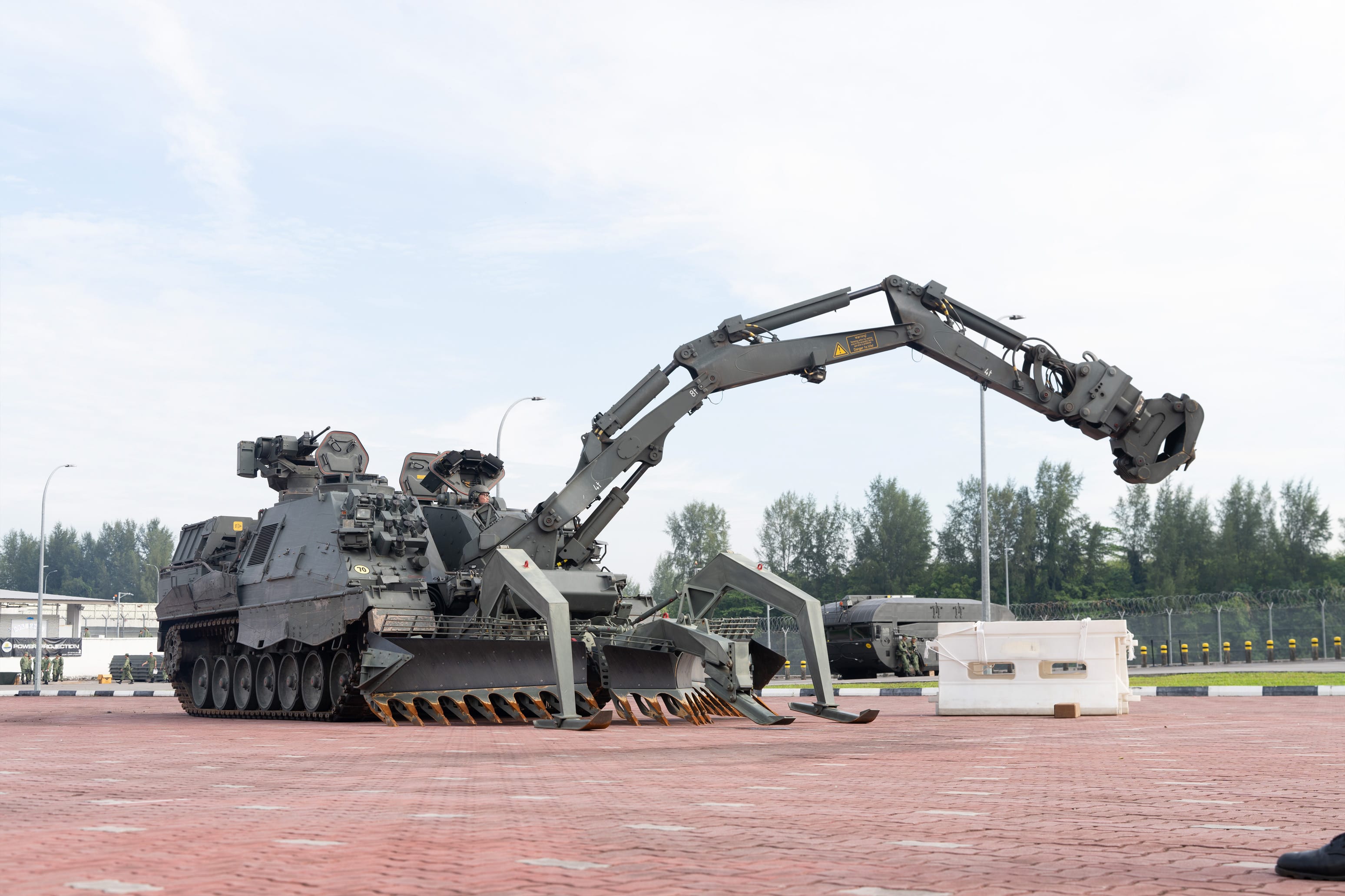 A military armored tracked vehicle with a large hydraulic crane arm and front dozer blade on a brick surface.