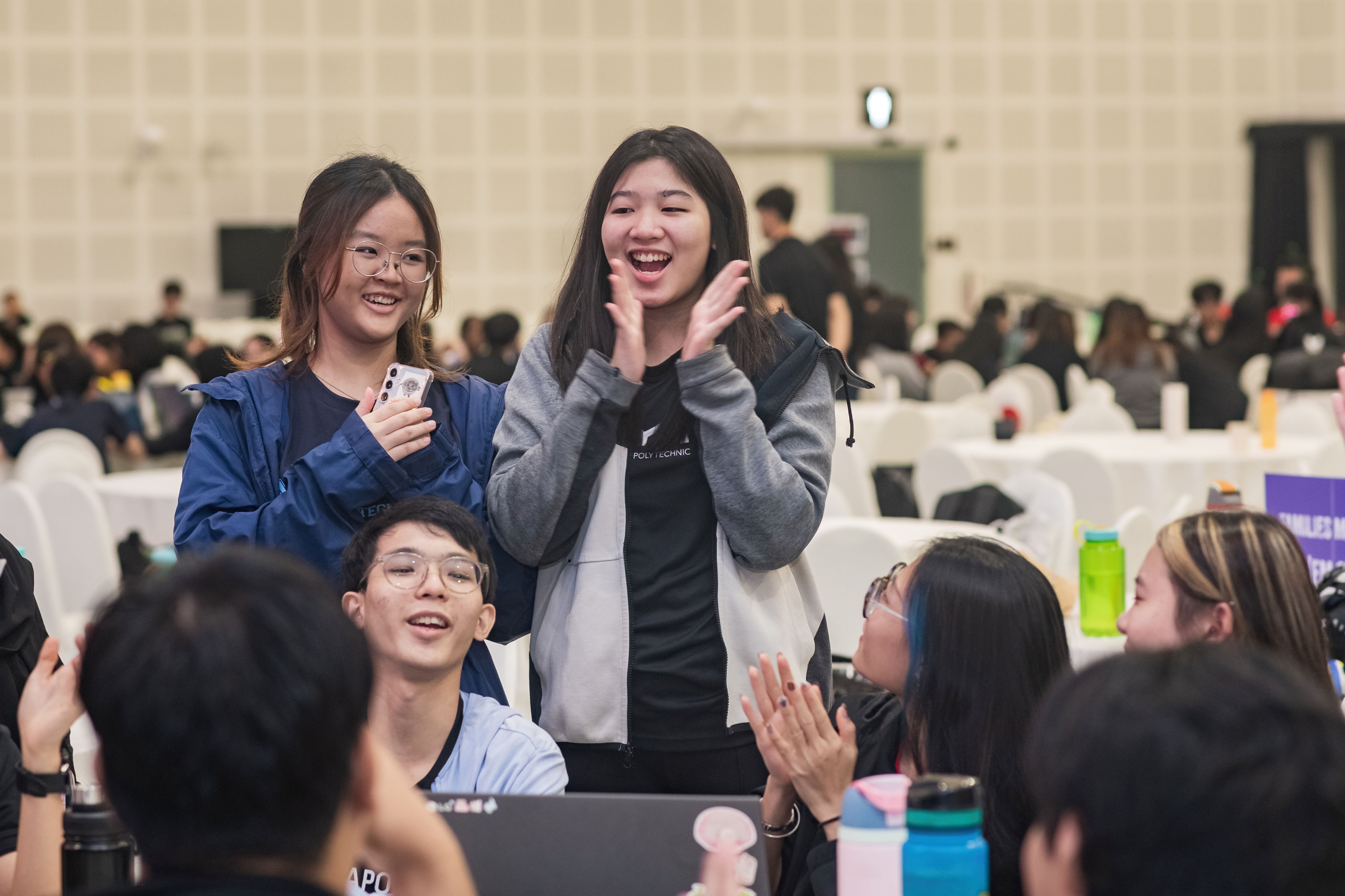 Young people smiling and clapping enthusiastically at a crowded indoor event with tables and chairs in the background.