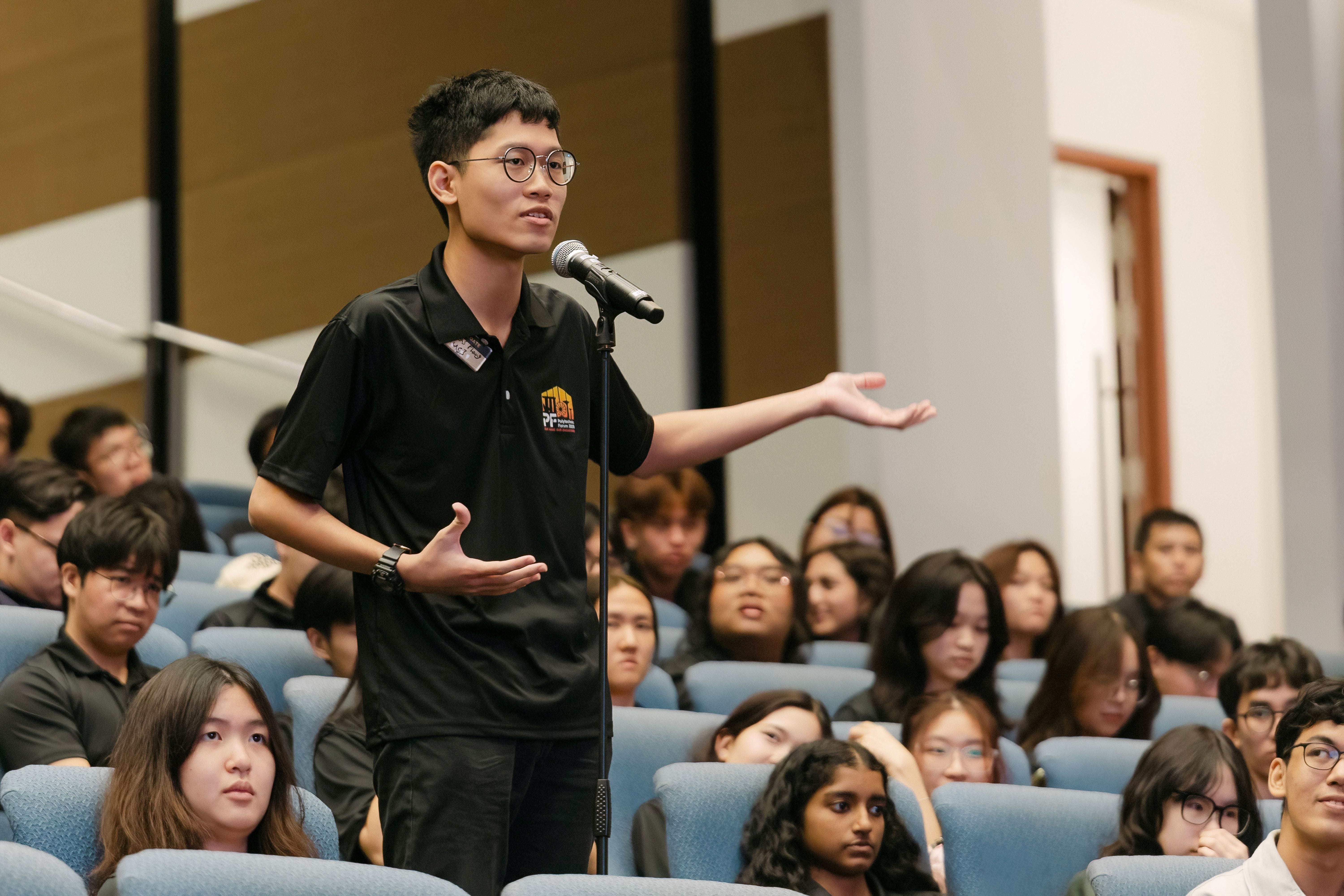 A young man with glasses speaks at a microphone stand in an auditorium filled with seated students.