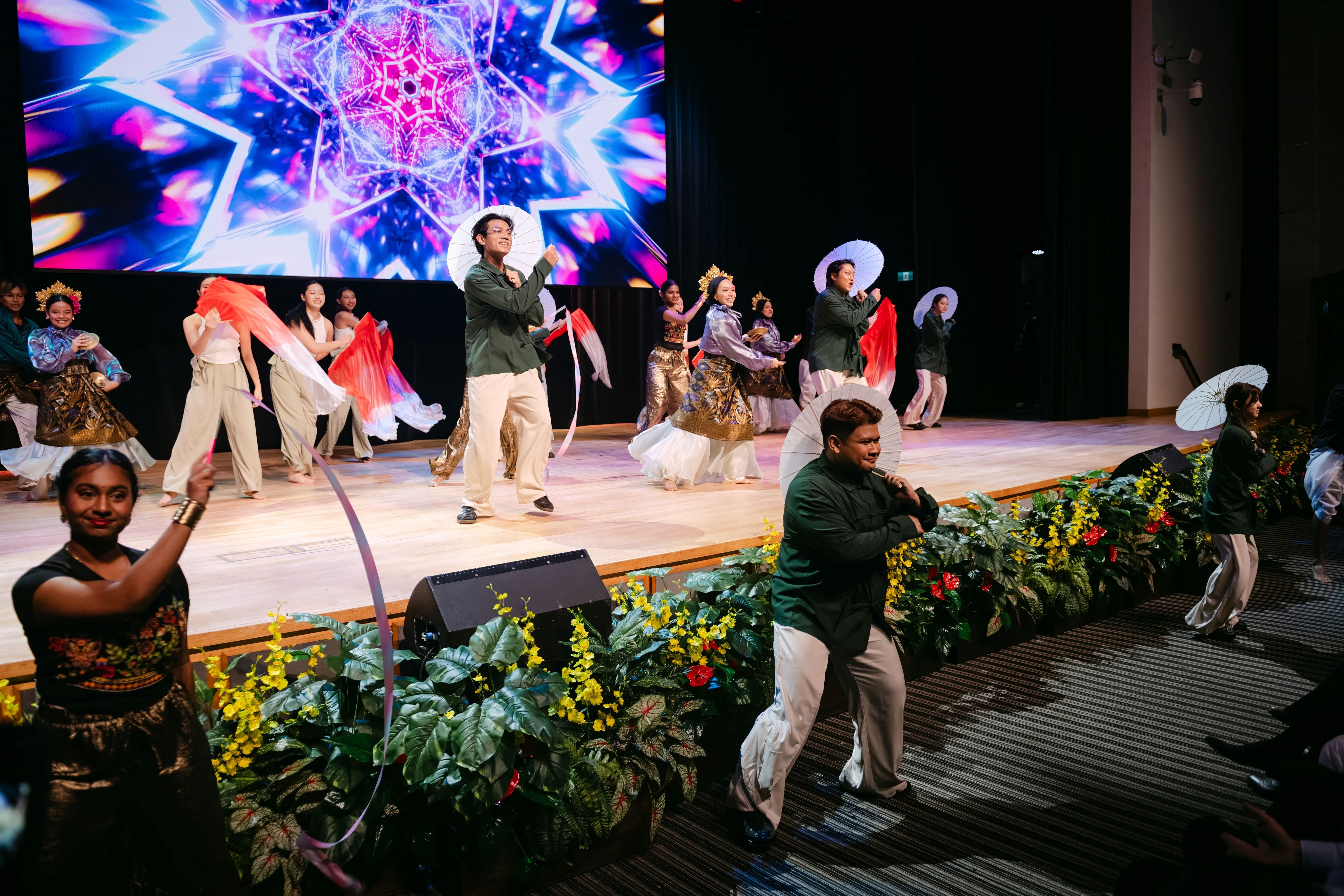 Performers in colorful costumes dance on a decorated stage with parasols and ribbons, backed by a vivid LED screen.