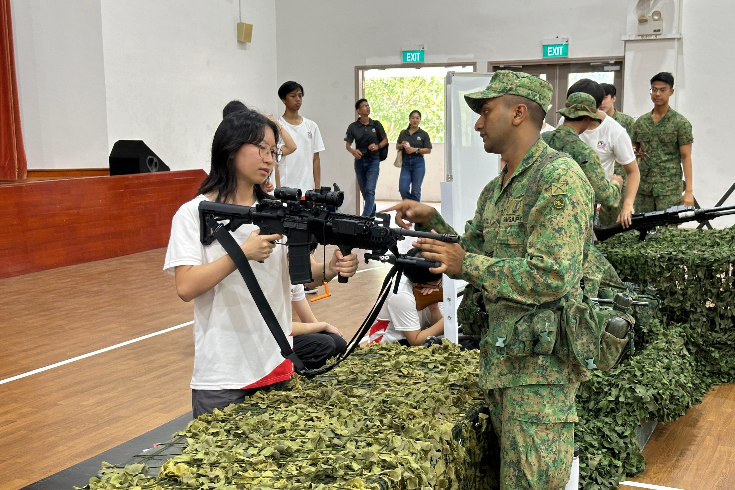 A soldier in camouflage uniform instructs a young woman holding a rifle at an indoor military display event.