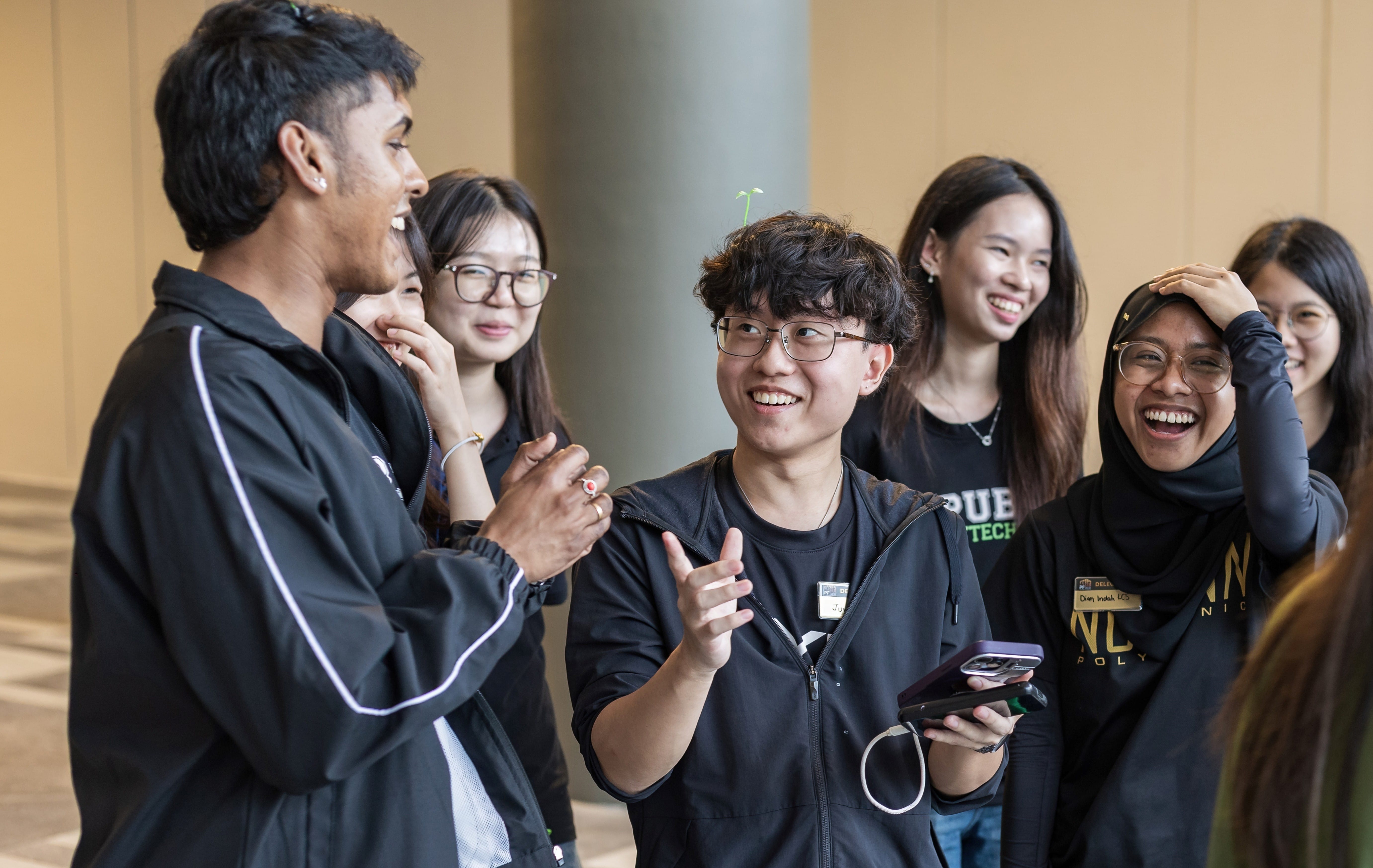 A group of smiling and laughing young students in black shirts talking together indoors.