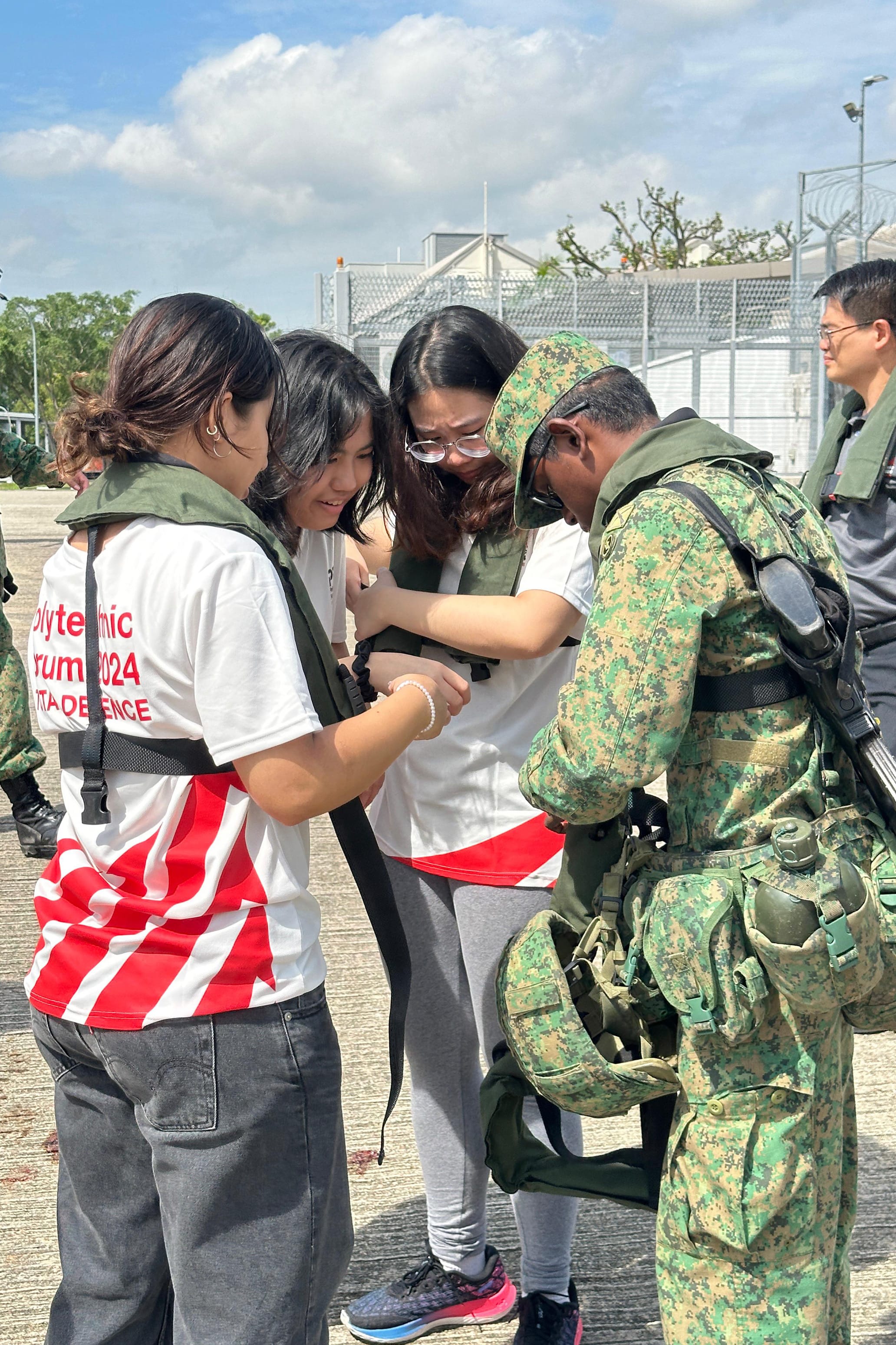A soldier in camouflage helps two young women adjust tactical vests outdoors at a 2024 polytechnic defence forum.